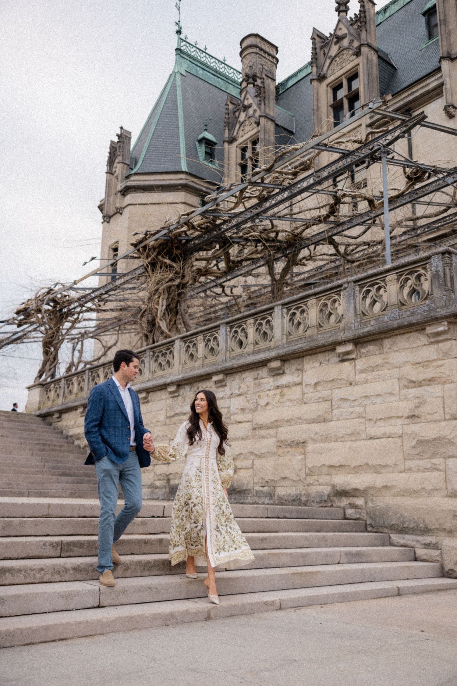 A couple walks down stone steps at the Biltmore Estate with ornate Gothic architecture and climbing vines in the background.