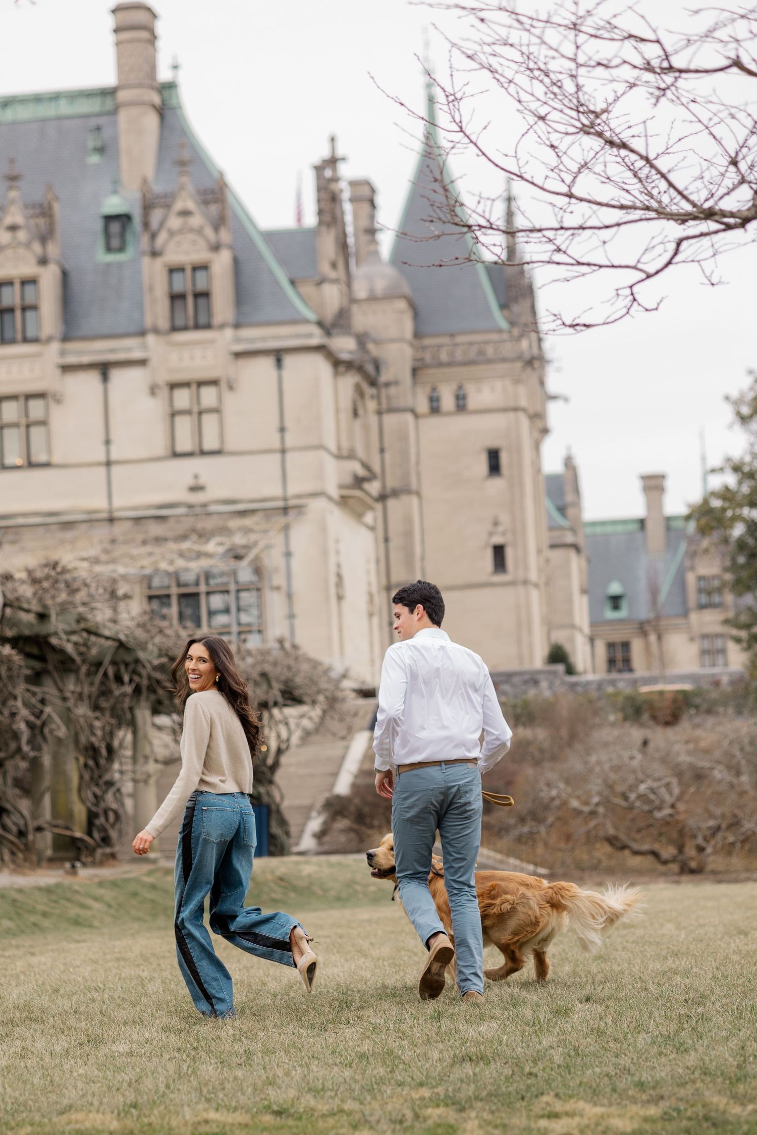 Two people walking with their golden retriever on the grounds of a majestic Gothic castle with turrets and spires.