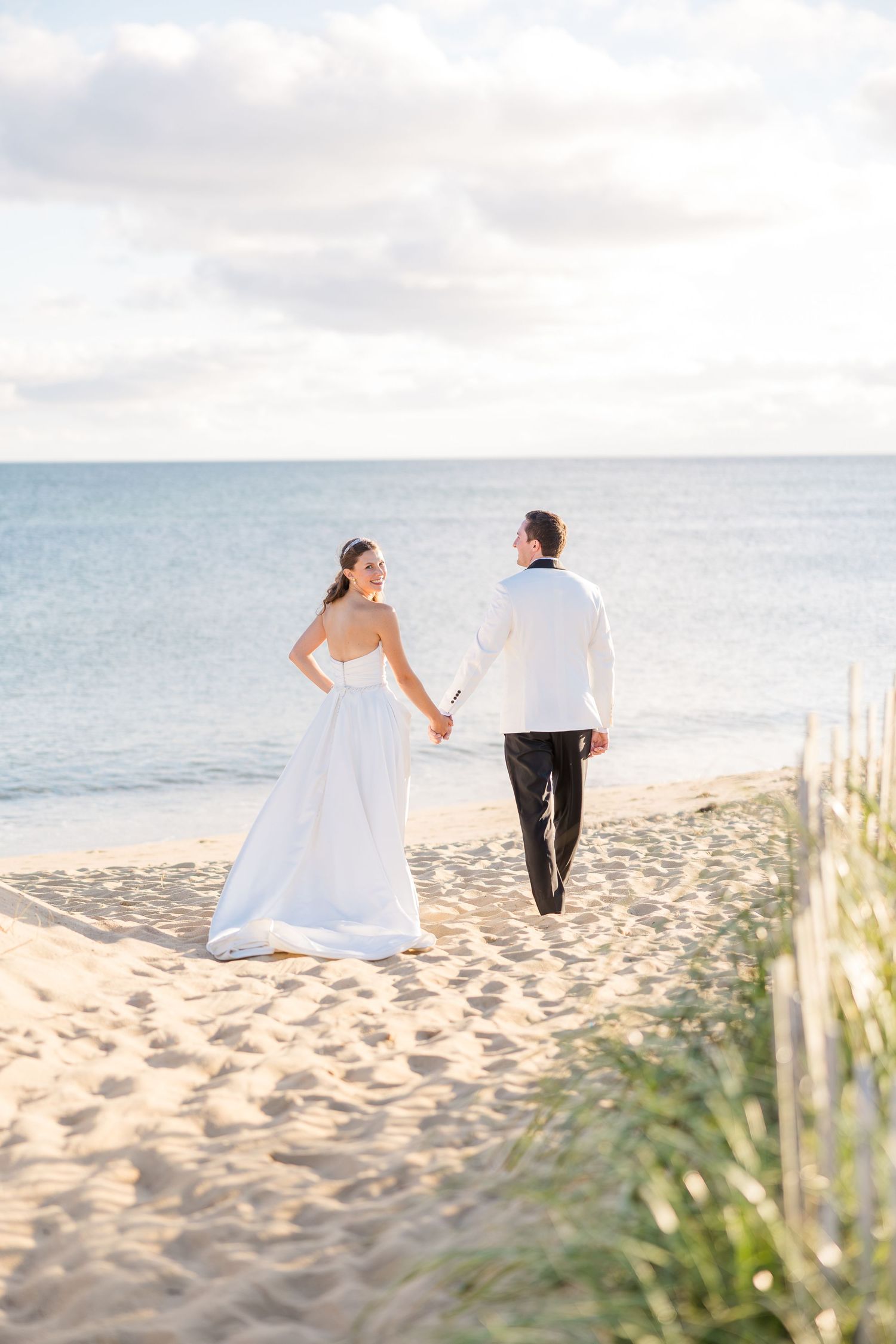Bride and groom walking hand in hand on a sandy beach toward the ocean in elegant white wedding attire.