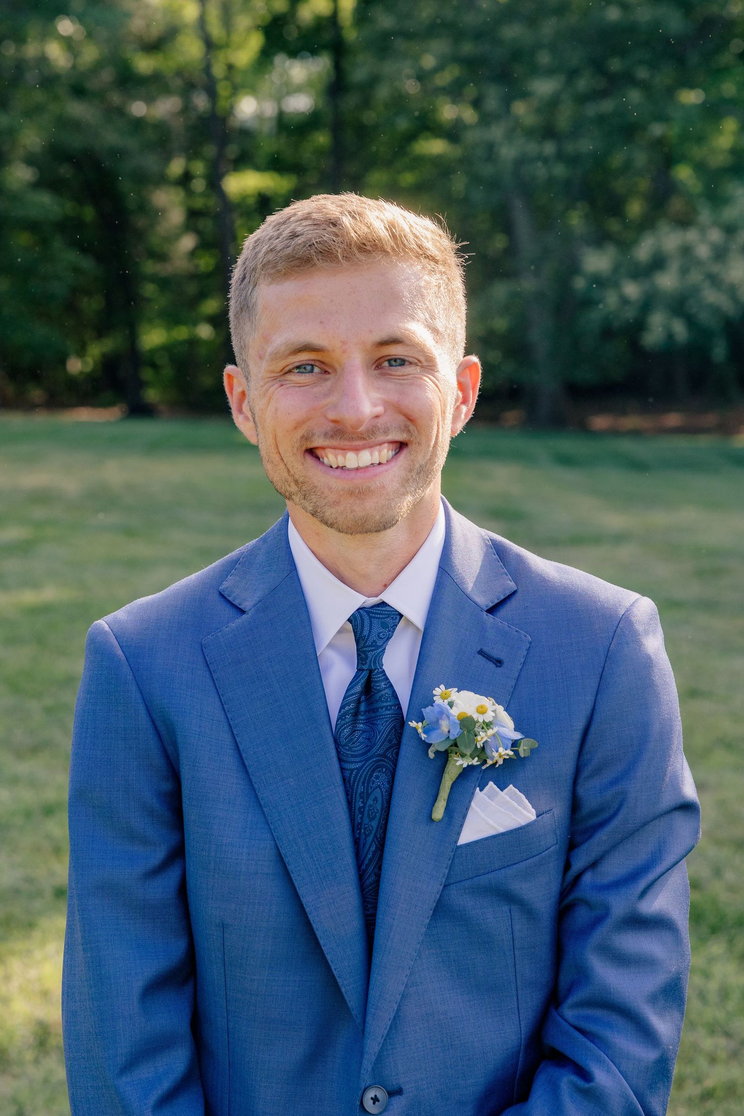 Smiling groom in blue suit with floral boutonniere stands outdoors on wedding day.