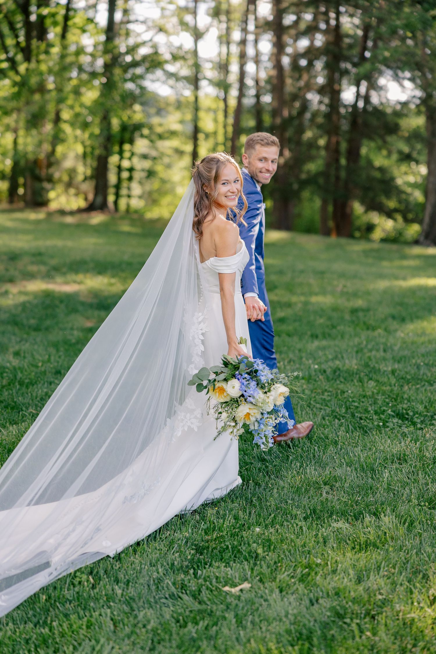 Bride and groom walking on lush green grass, bride wearing flowing veil and holding blue and yellow bouquet.