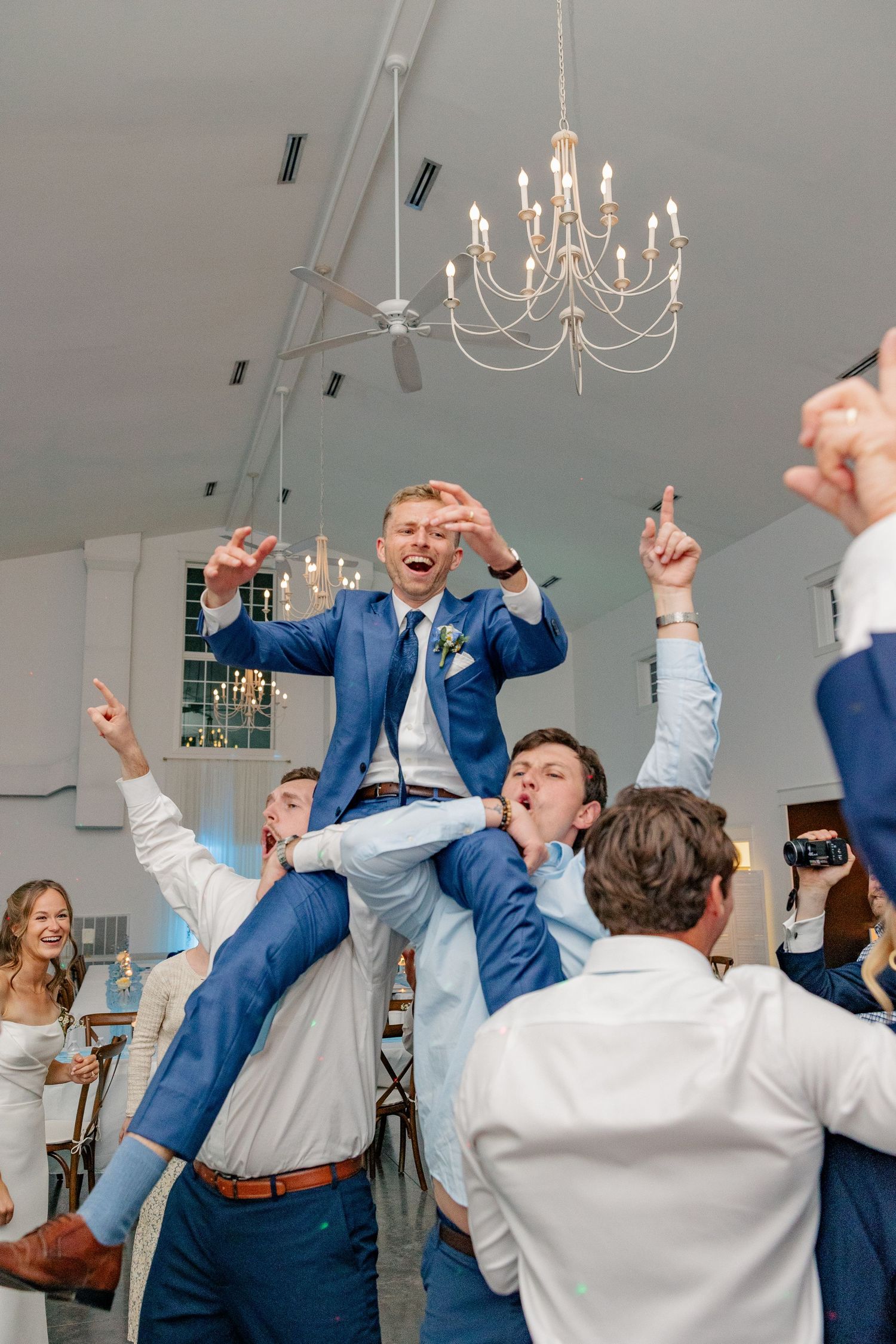 Groom in blue suit lifted by groomsmen celebrating on dance floor under elegant chandelier at wedding reception.