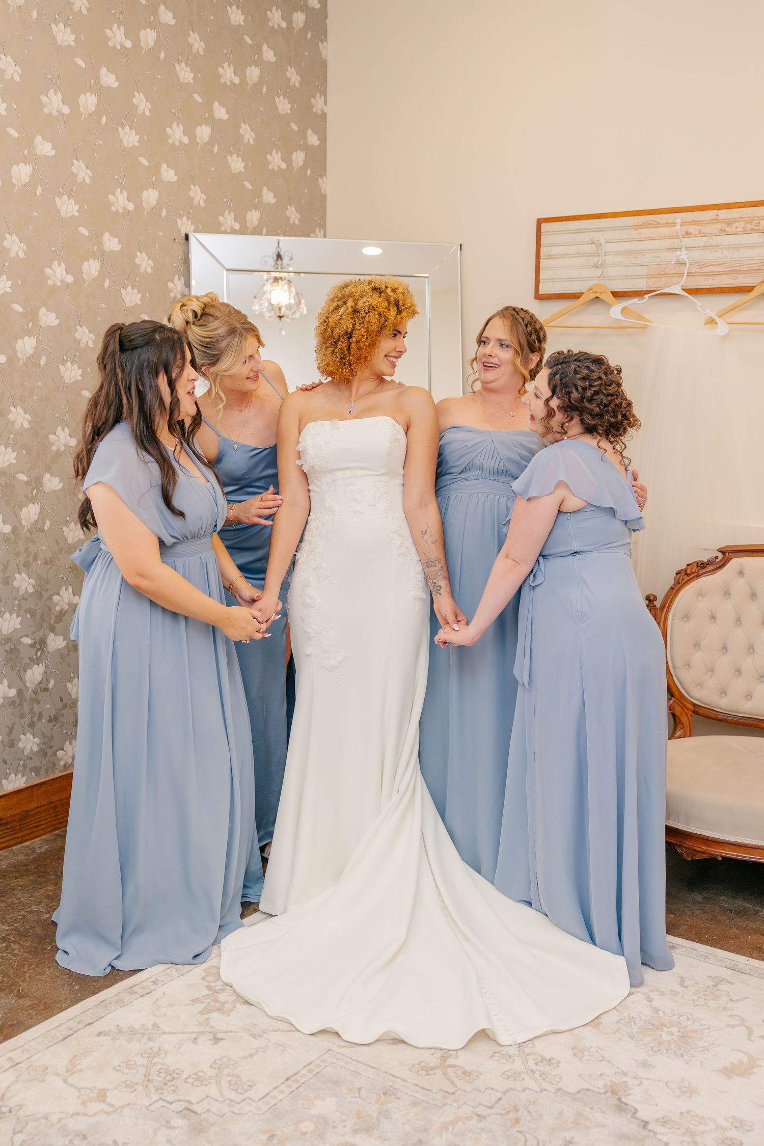 Bride in white gown surrounded by four bridesmaids in dusty blue dresses in elegant bridal suite.