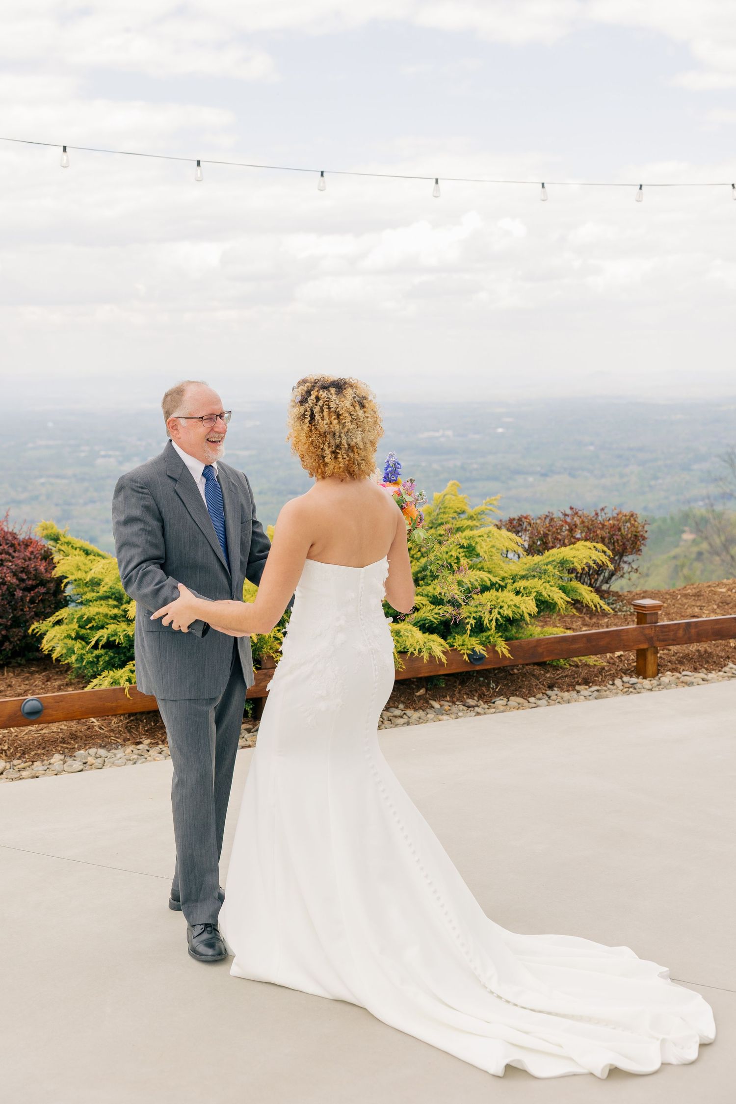 Bride in strapless gown shares a first look moment with her father on an outdoor terrace with scenic views.