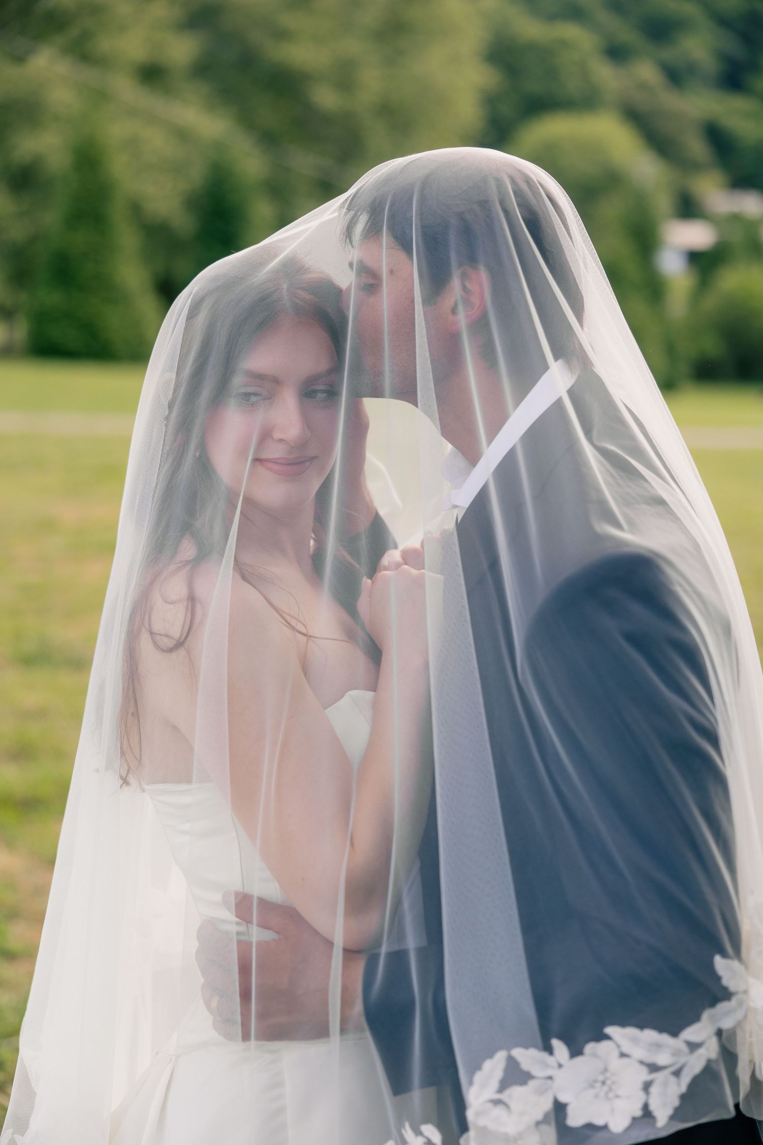 Romantic bride and groom share a kiss hidden beneath a delicate lace-trimmed wedding veil outdoors.