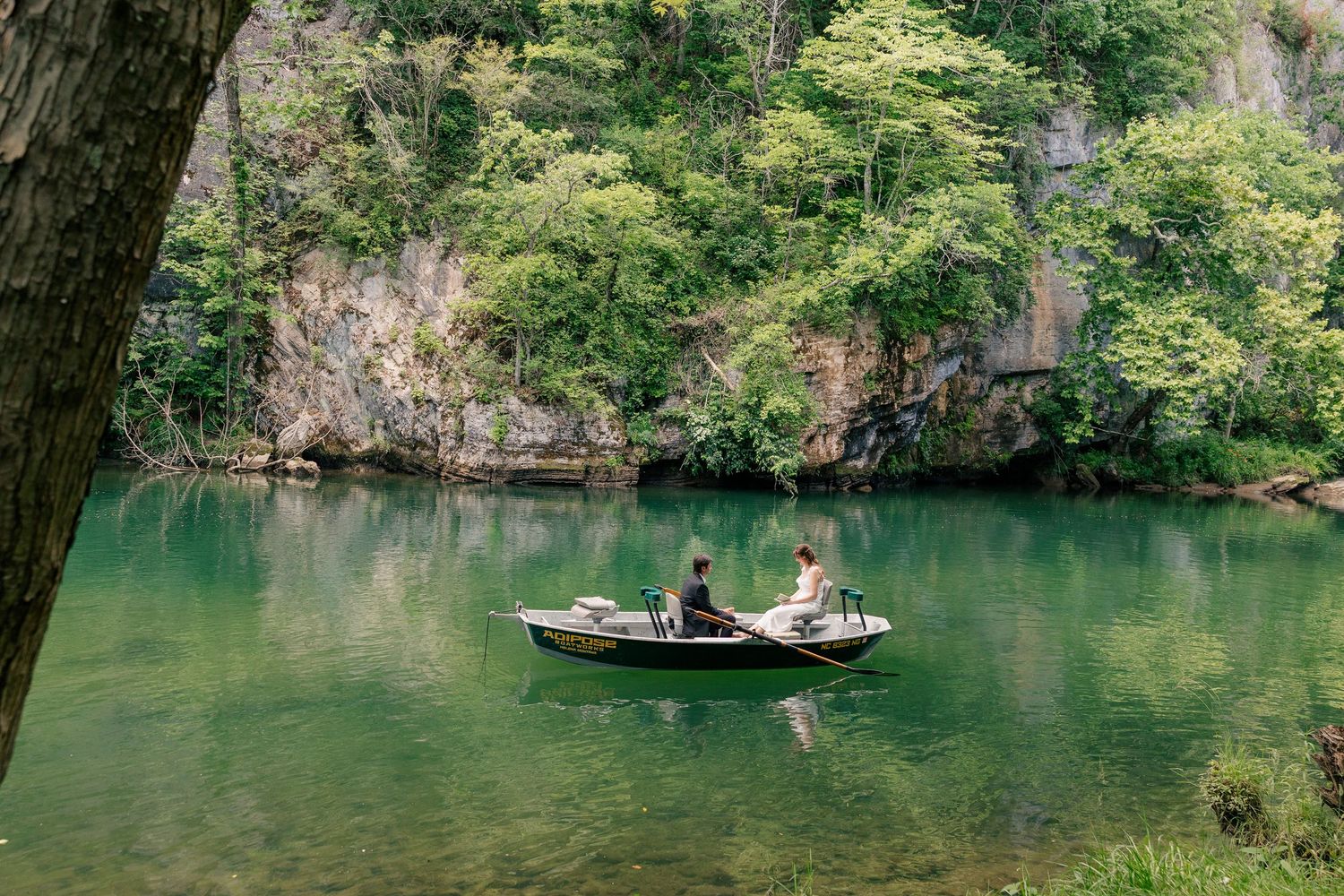 Two people sit in a small wooden boat on a serene green lake surrounded by rocky cliffs and trees.