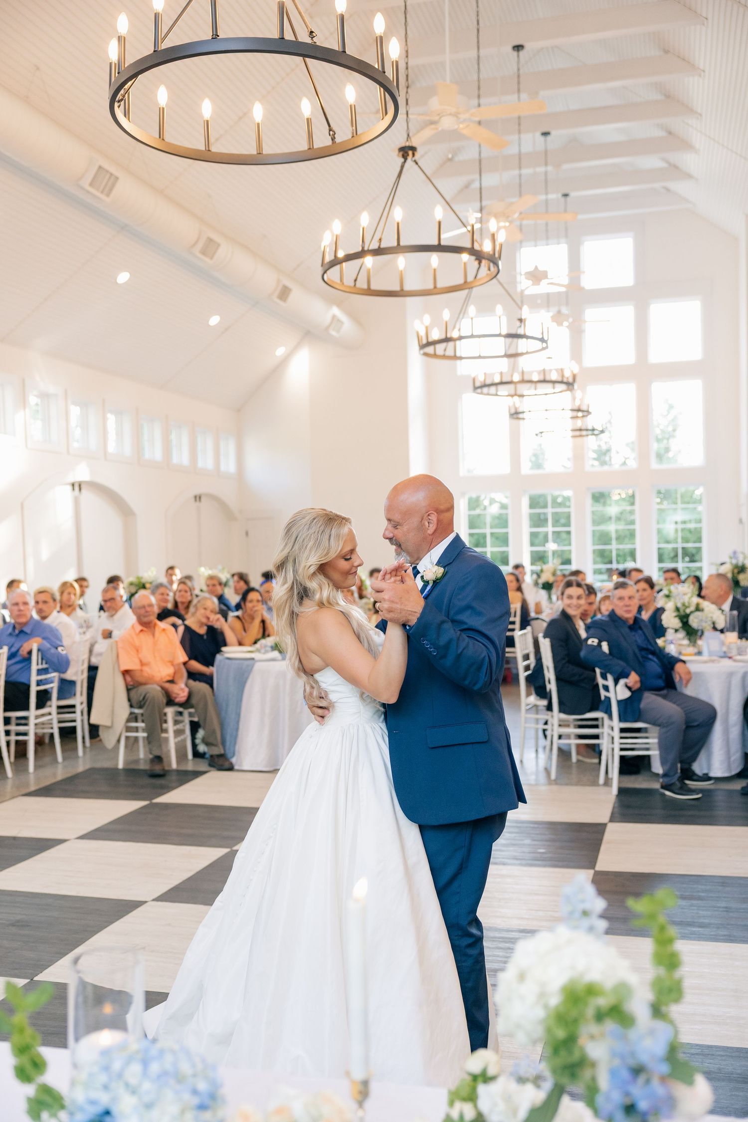 Bride and groom share first dance in elegant white venue with chandeliers and guests seated at tables.