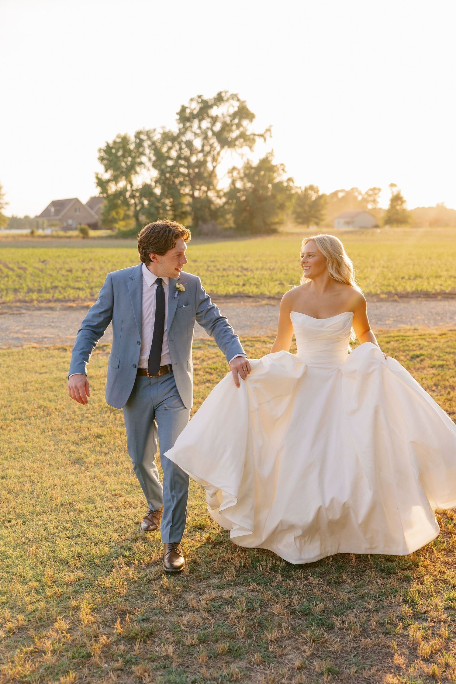 Bride and groom laugh and run through a sunlit field at golden hour during wedding portraits.