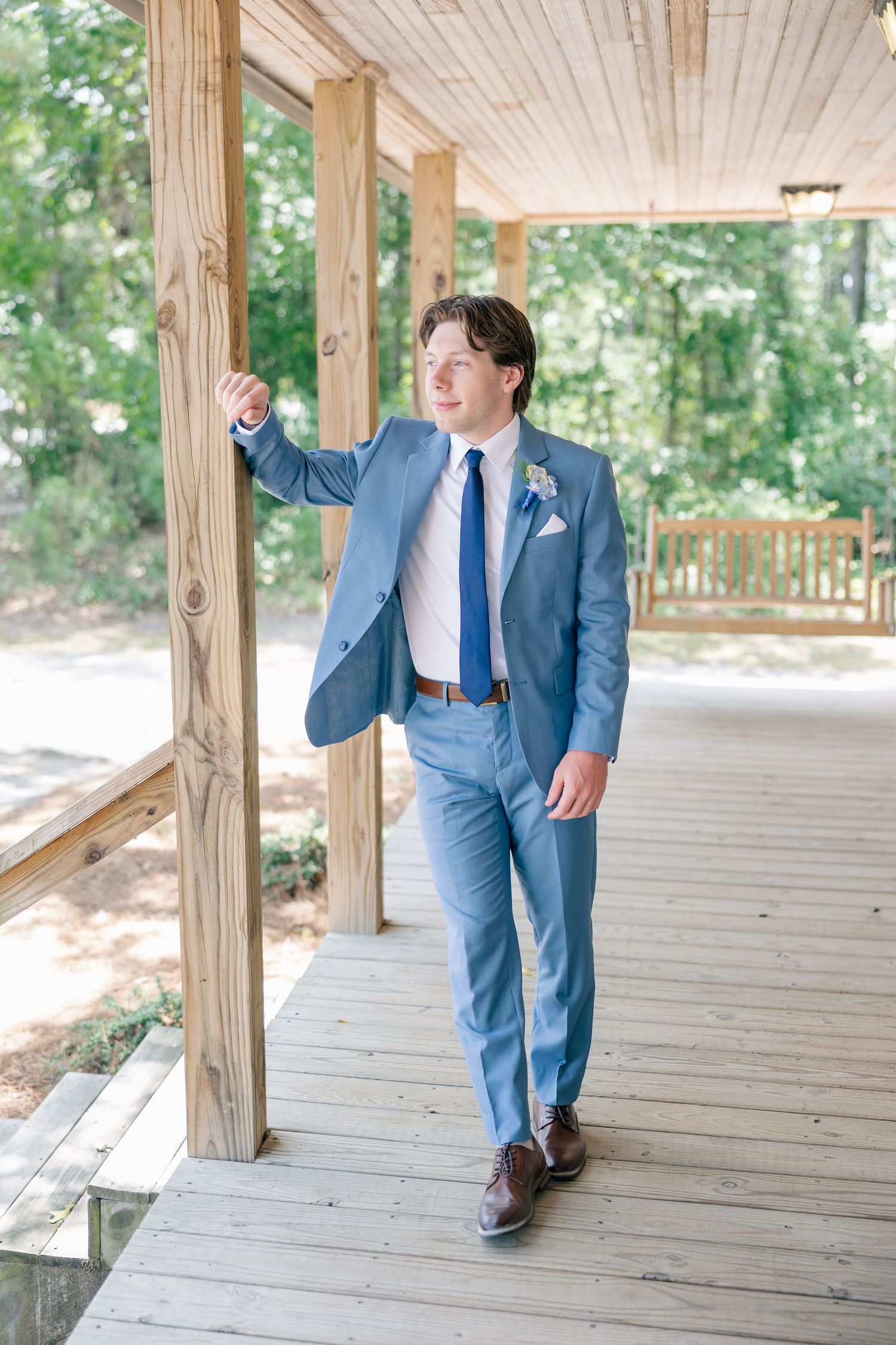 Dapper groom in light blue suit with boutonniere poses on wooden deck leaning against timber post.
