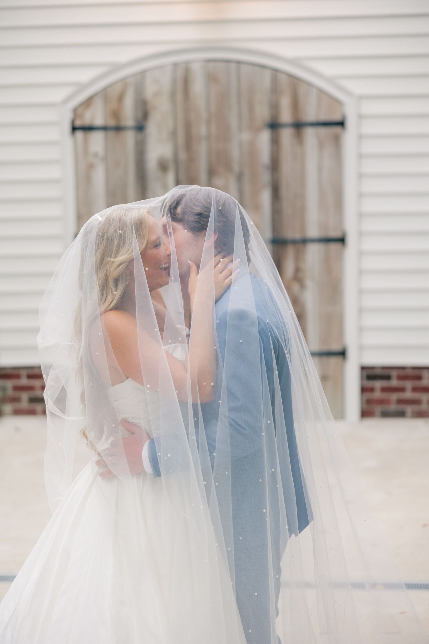 Bride and groom share romantic kiss hidden under pearl-adorned wedding veil outside barn venue.