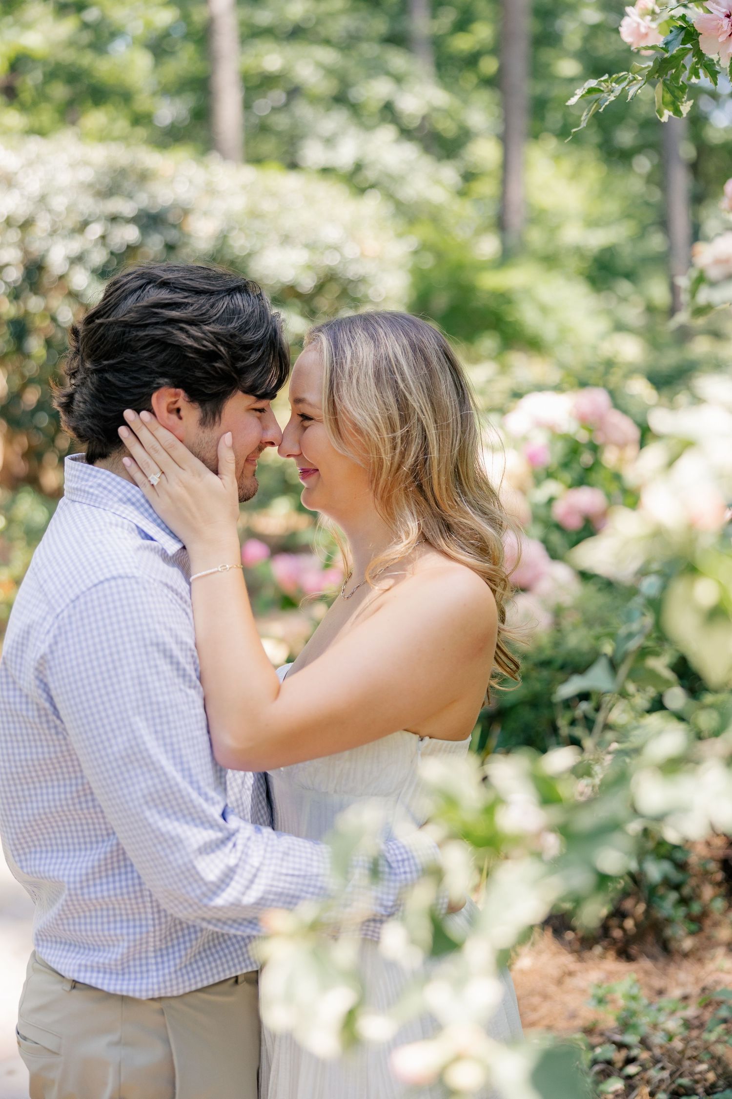 A couple shares a romantic moment in a lush garden surrounded by blooming pink flowers and greenery.