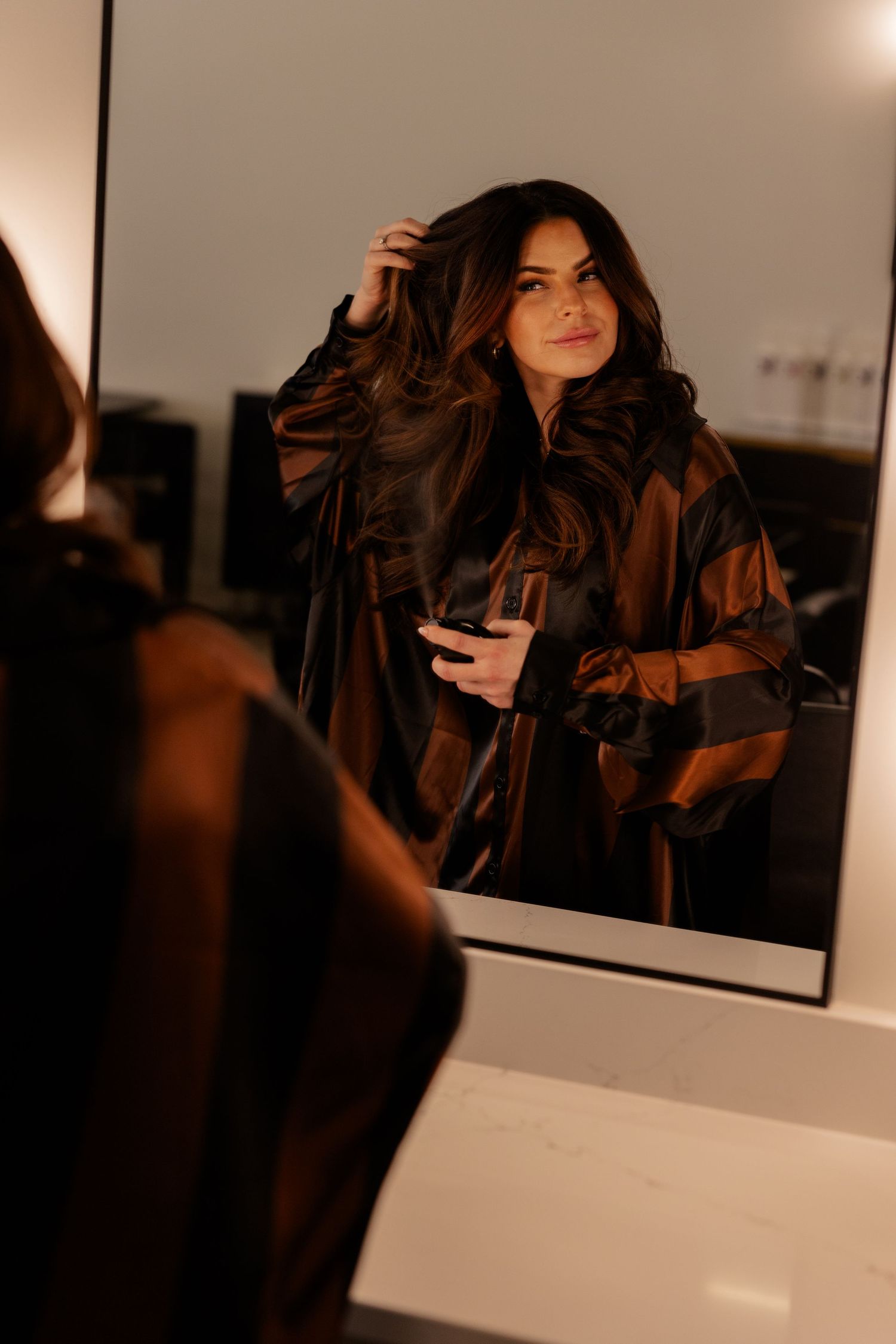 A reflection in a mirror shows someone with long dark wavy hair holding a phone in warm moody lighting.