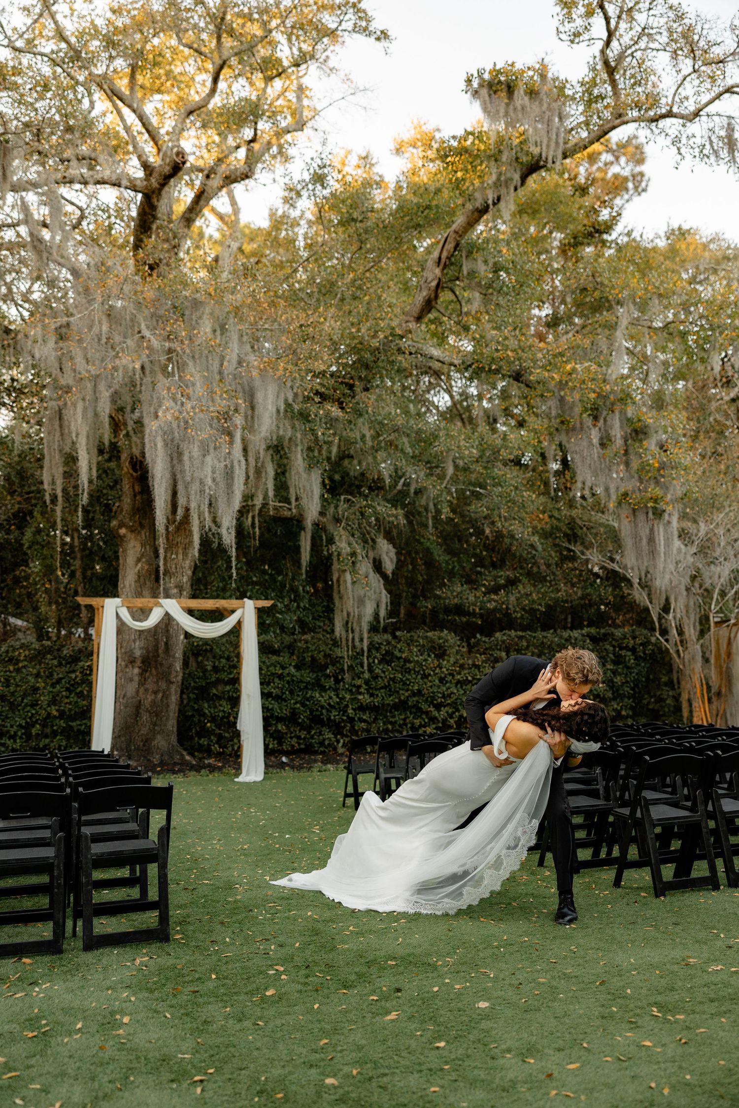 An outdoor wedding ceremony setup under Spanish moss draped trees with white chairs and arch decor.