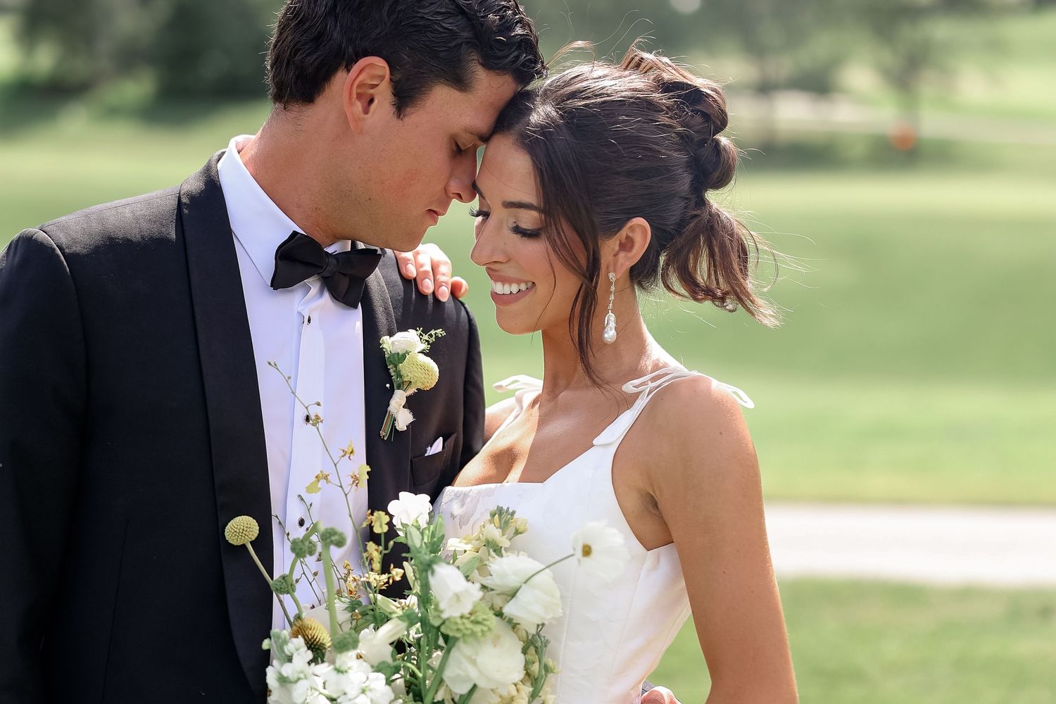 Bride and groom share intimate moment, she holds white bouquet in elegant wedding attire on lush green golf course.
