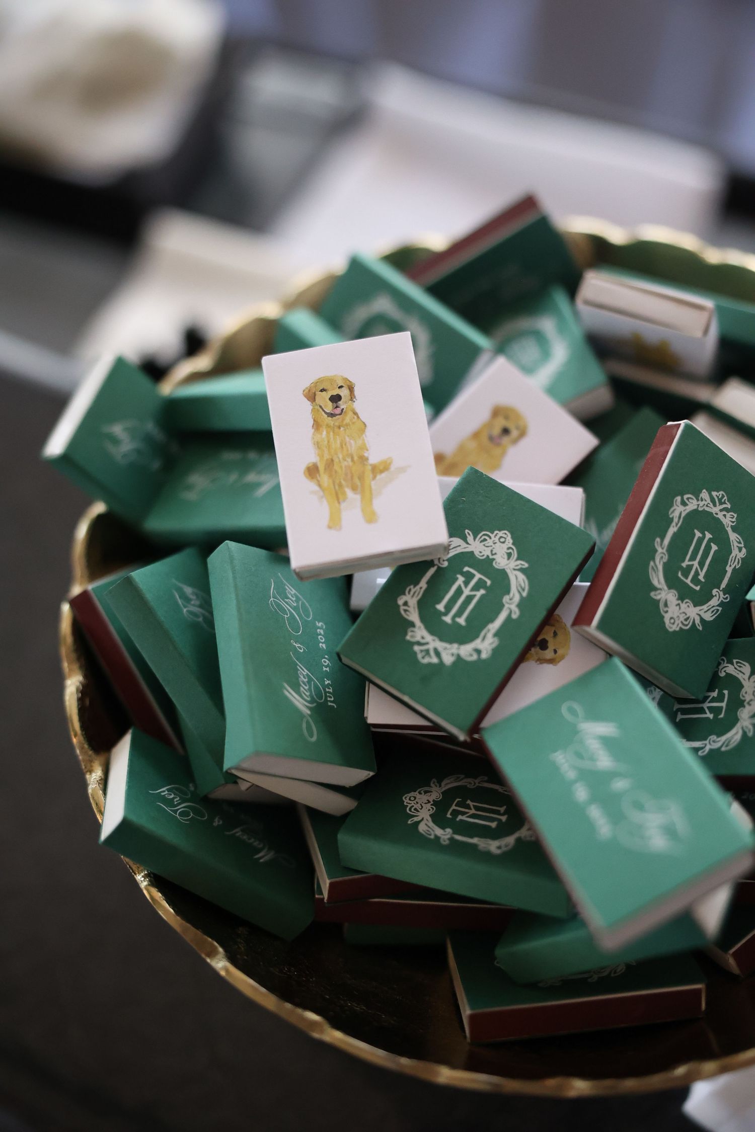 A gold bowl filled with green personalized matchboxes featuring monogram designs and a golden retriever illustration for a wedding.