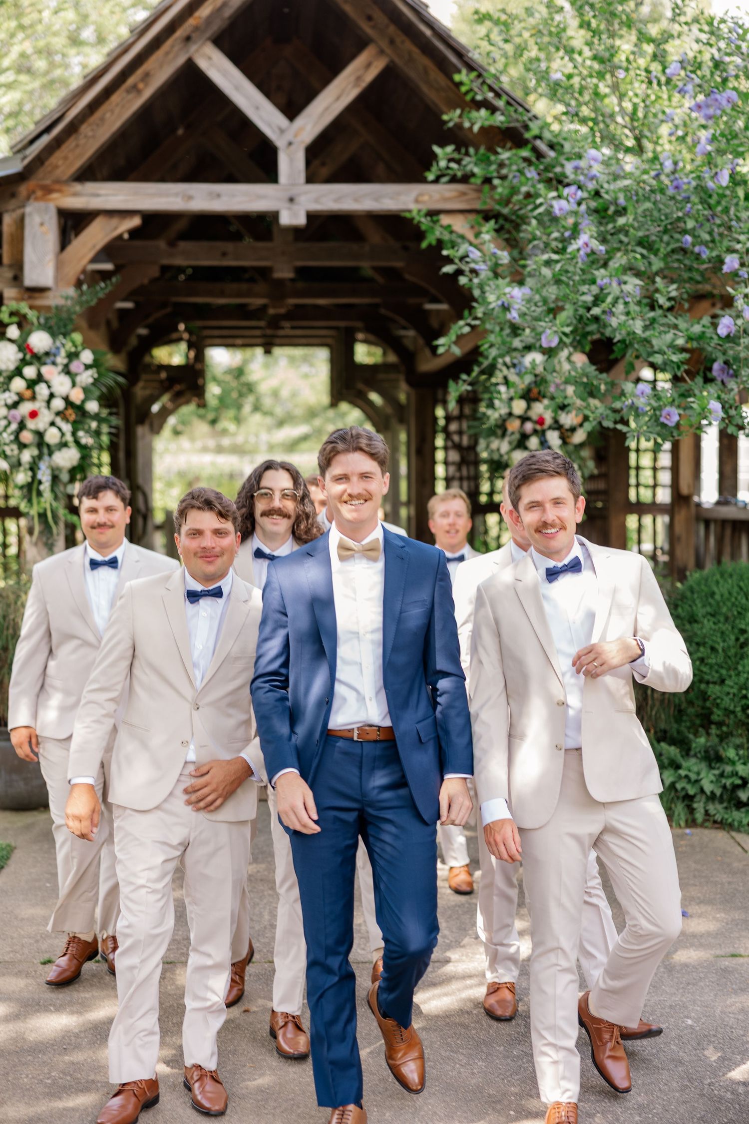 Wedding party poses outdoors with groomsmen in beige suits and navy blue accents under a rustic wooden pavilion.