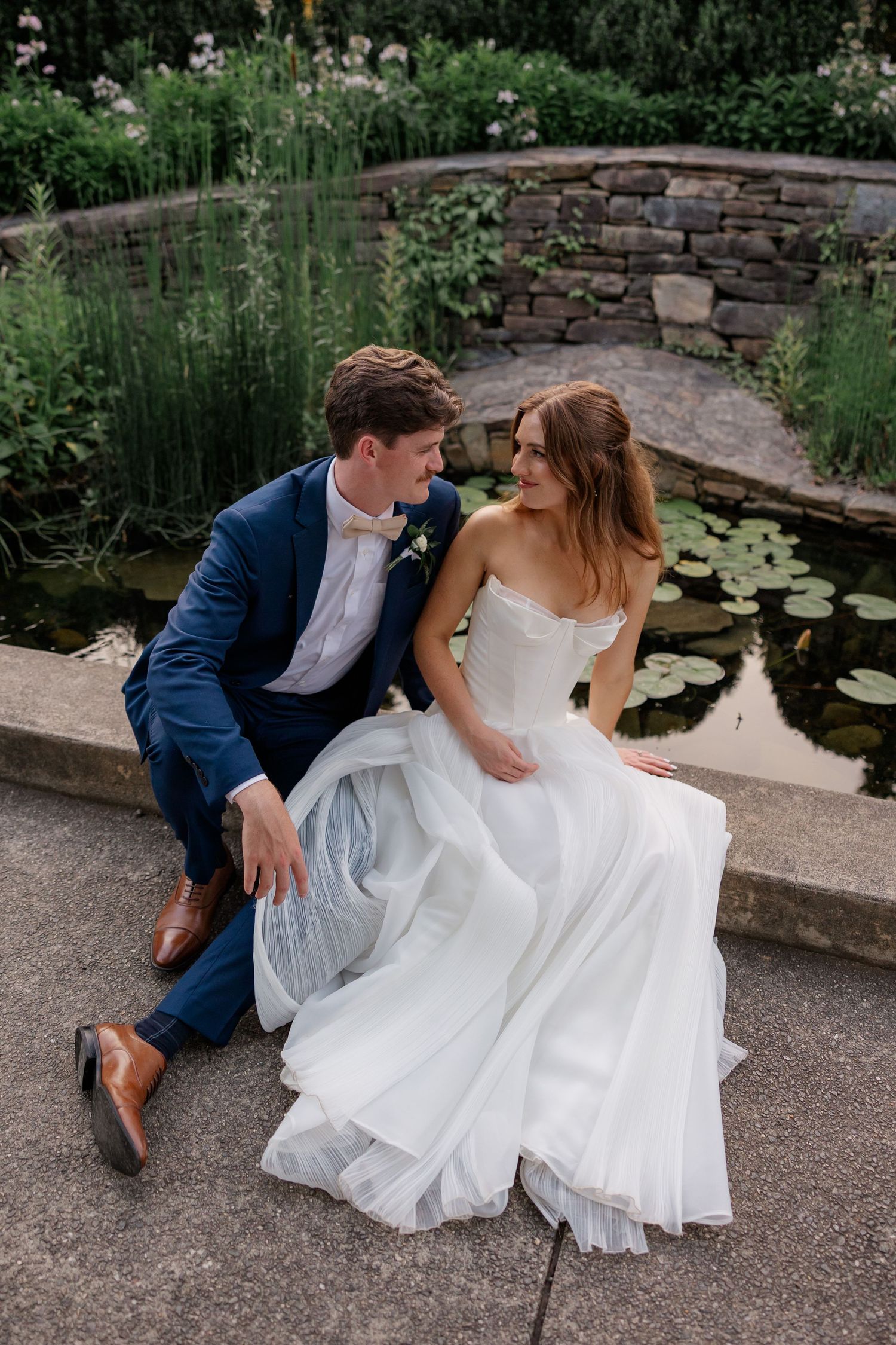 A couple in wedding attire sits by a stone wall and lily pond in a romantic garden setting.