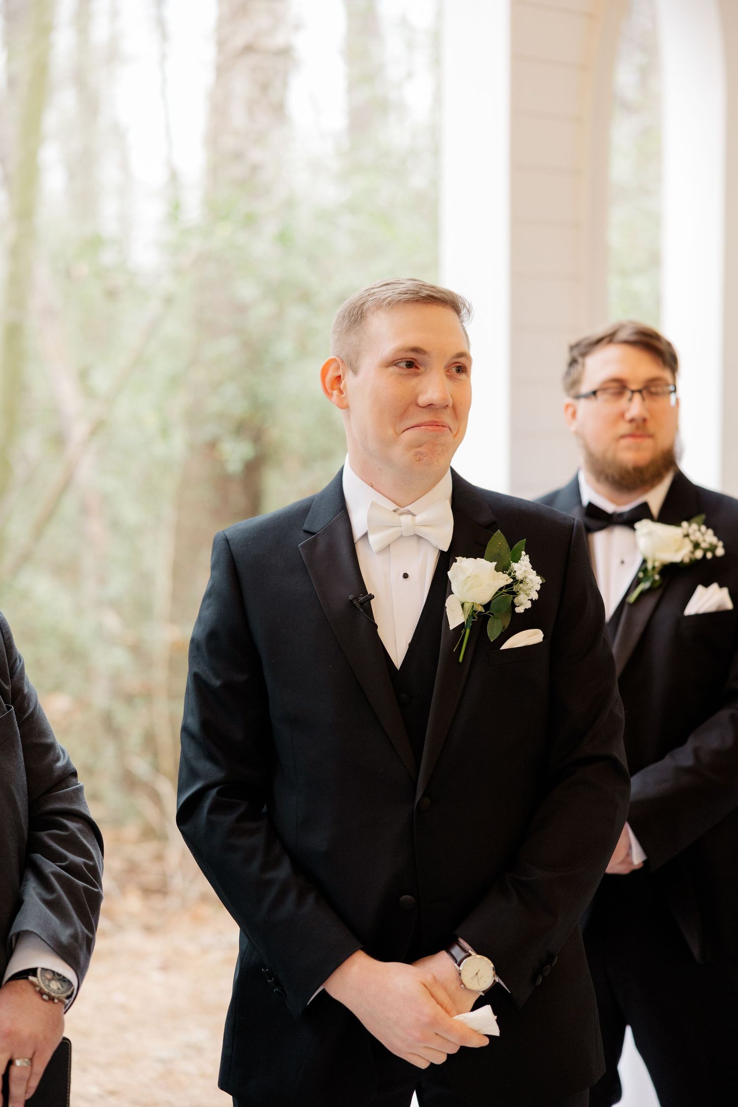Groomsmen in black tuxedos with white boutonnieres stand in line at wedding ceremony.