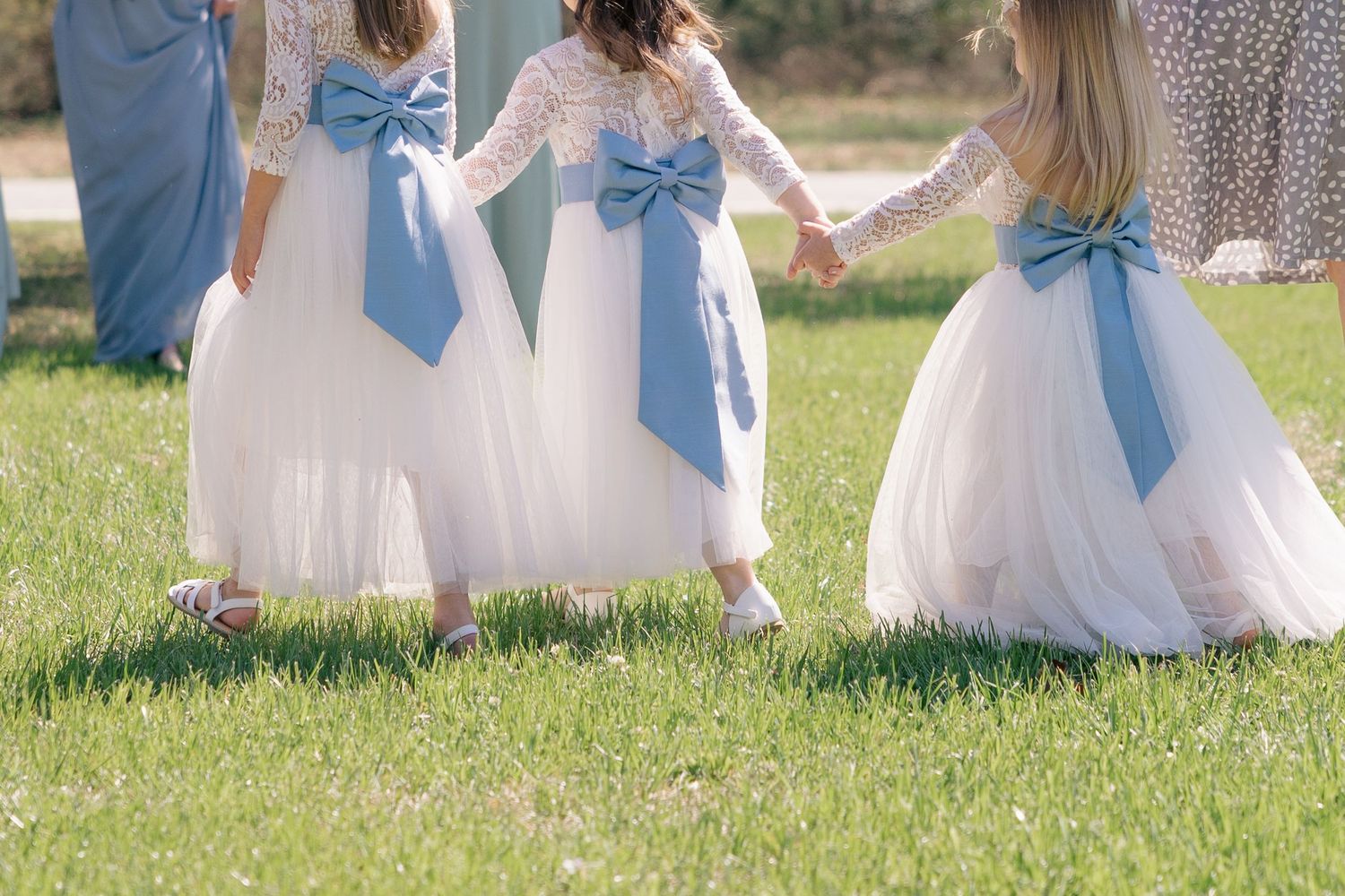 Bridesmaids in white dresses with light blue bows sitting together on grass outdoors.