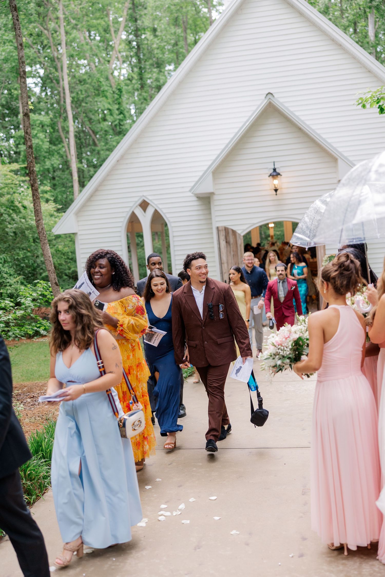 Wedding party exits charming white chapel while guests line walkway in blue and pink dresses.