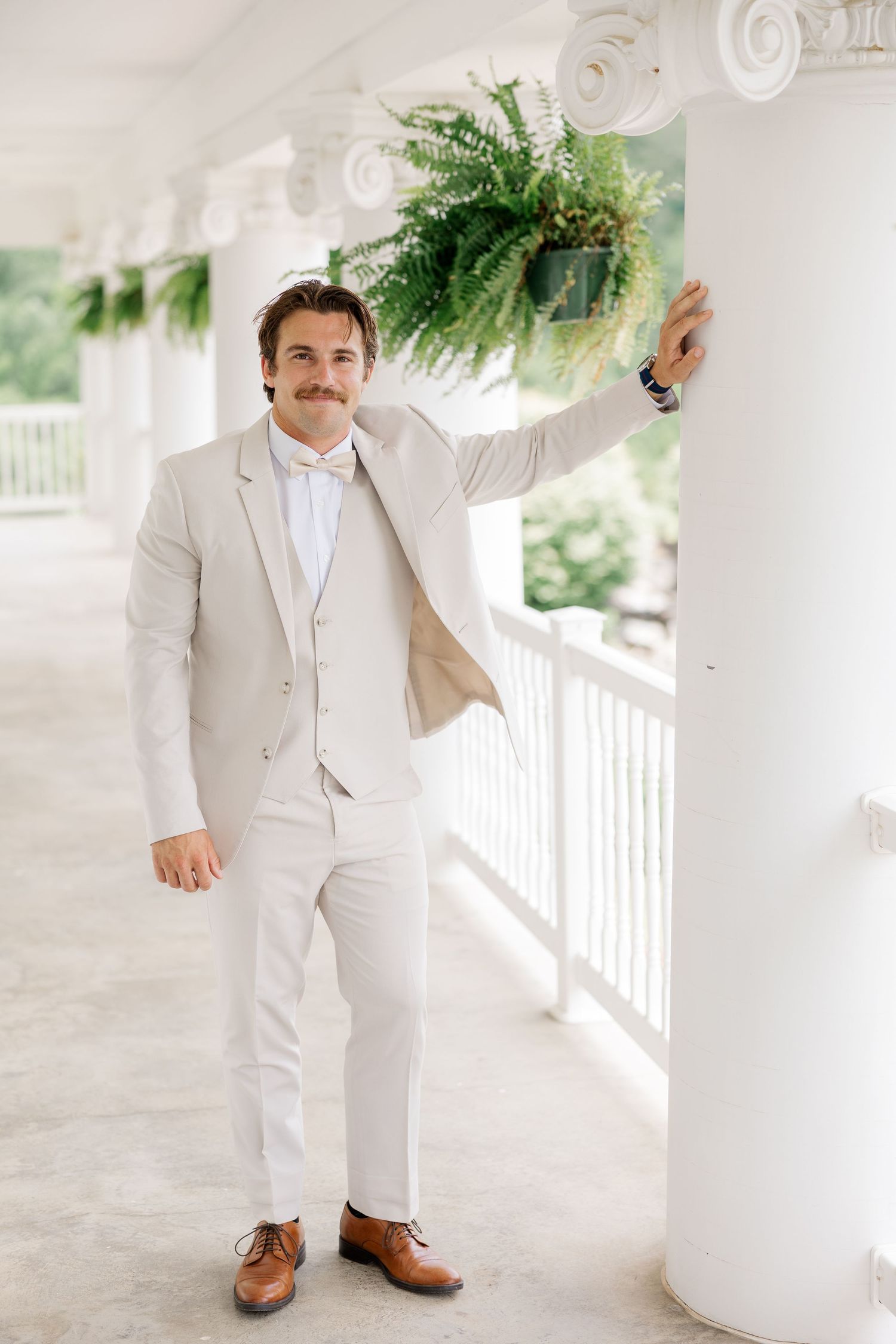 A young child in a light beige suit and brown shoes stands on a white porch.