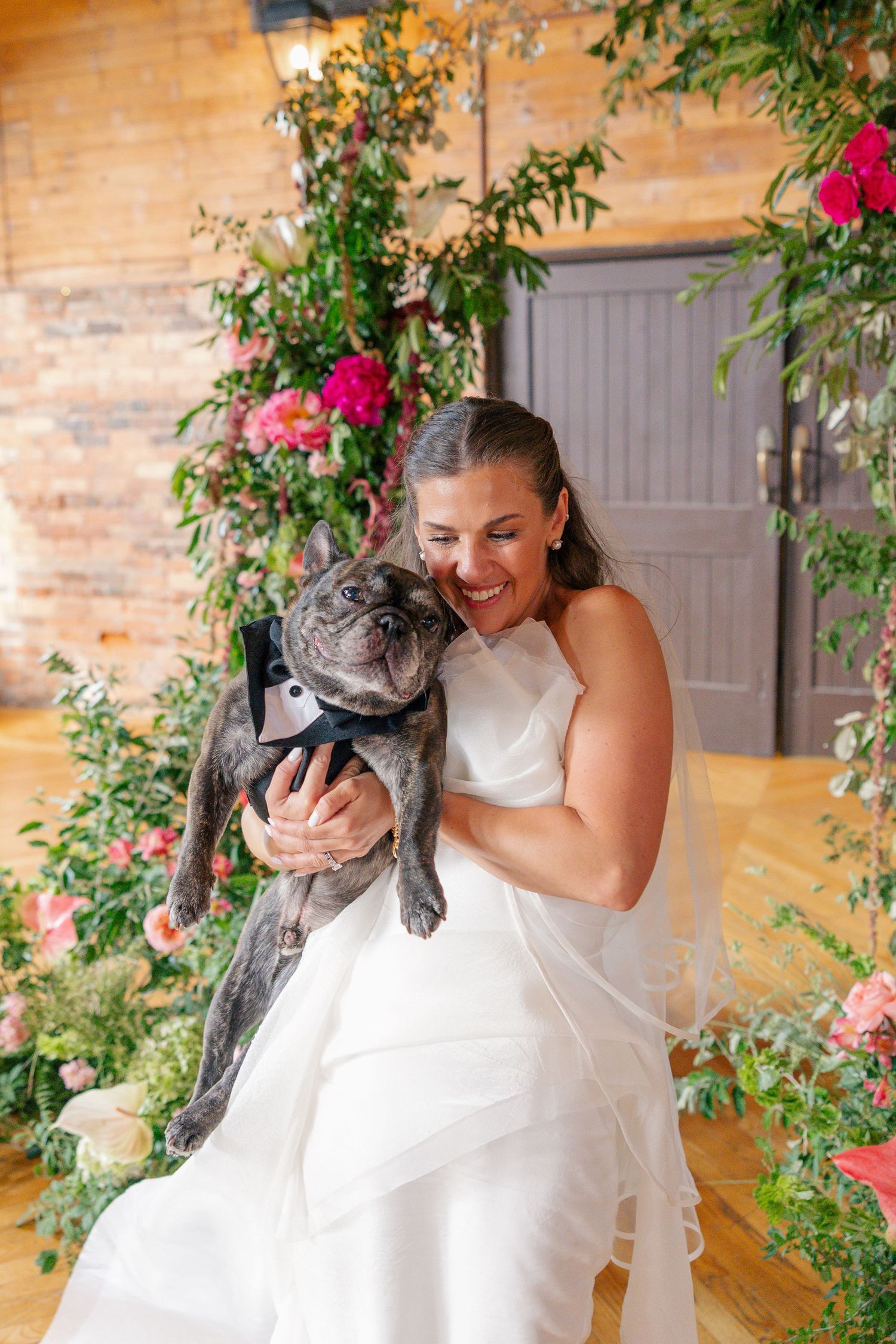 Joyful bride in white gown cuddles French bulldog in tiny tuxedo amid pink floral backdrop.