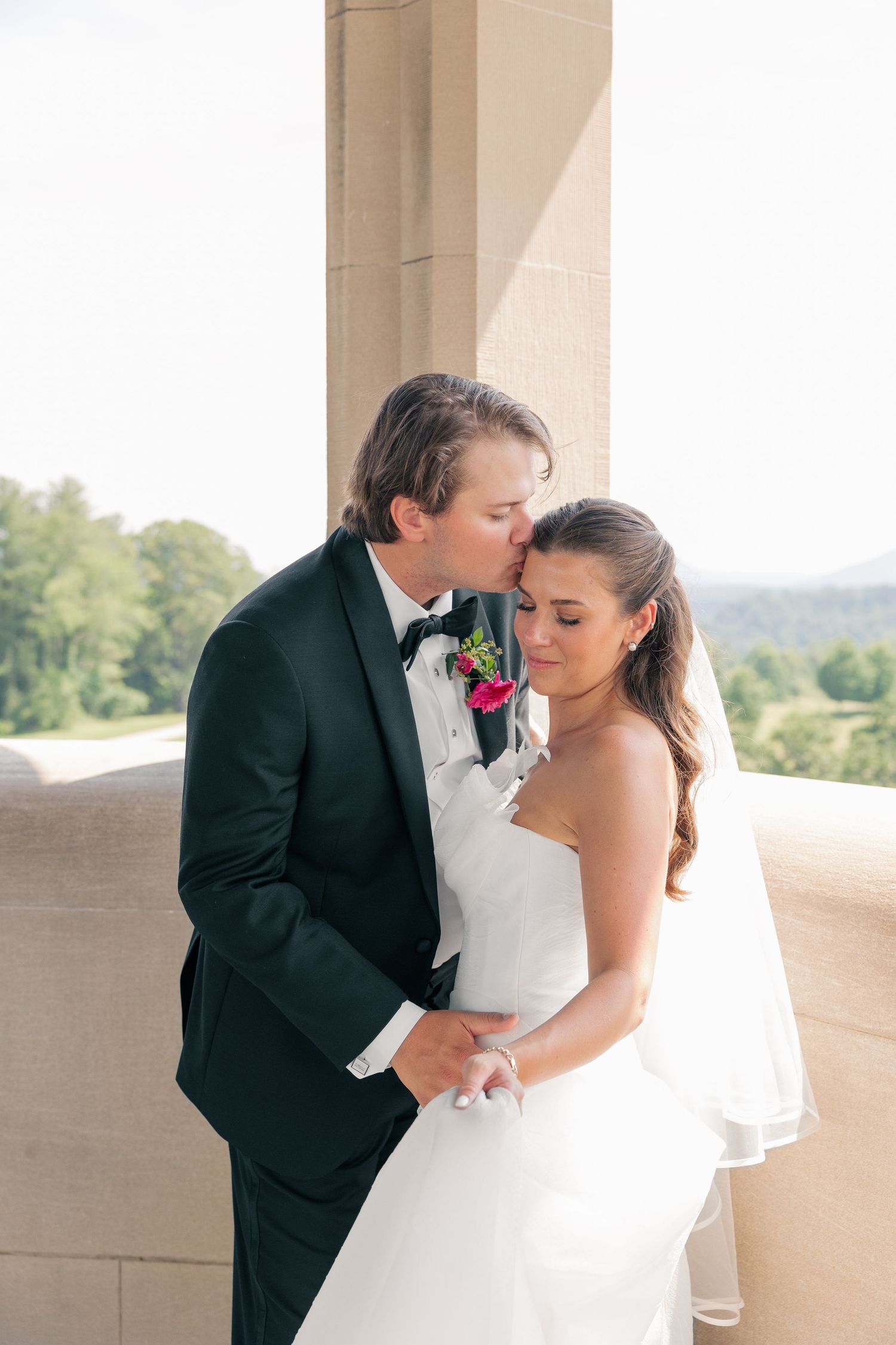 Groom tenderly kisses bride on forehead between stone columns with lush landscape backdrop.