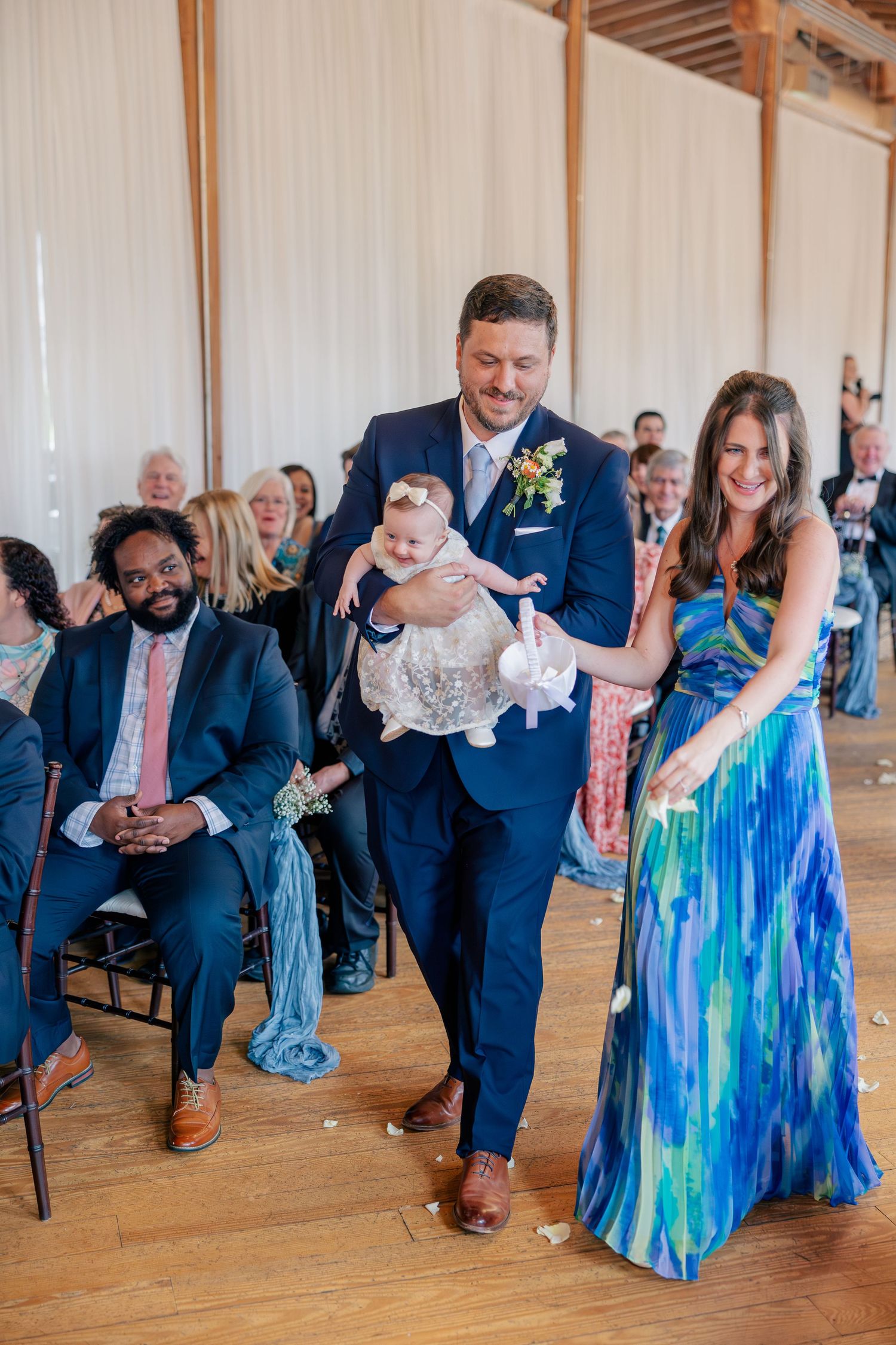 Groomsman carries a baby flower girl down the aisle during a bright indoor wedding ceremony.