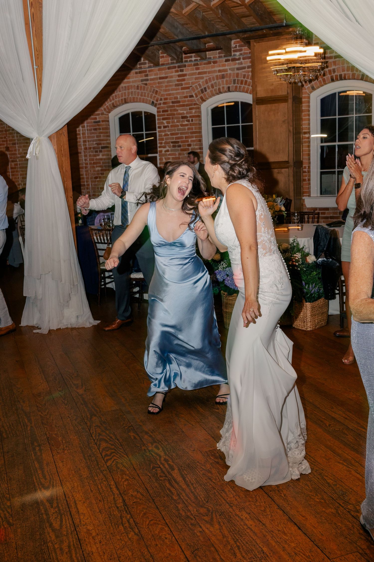 Bridesmaid in blue satin gown laughs with bride while dancing at wedding reception.