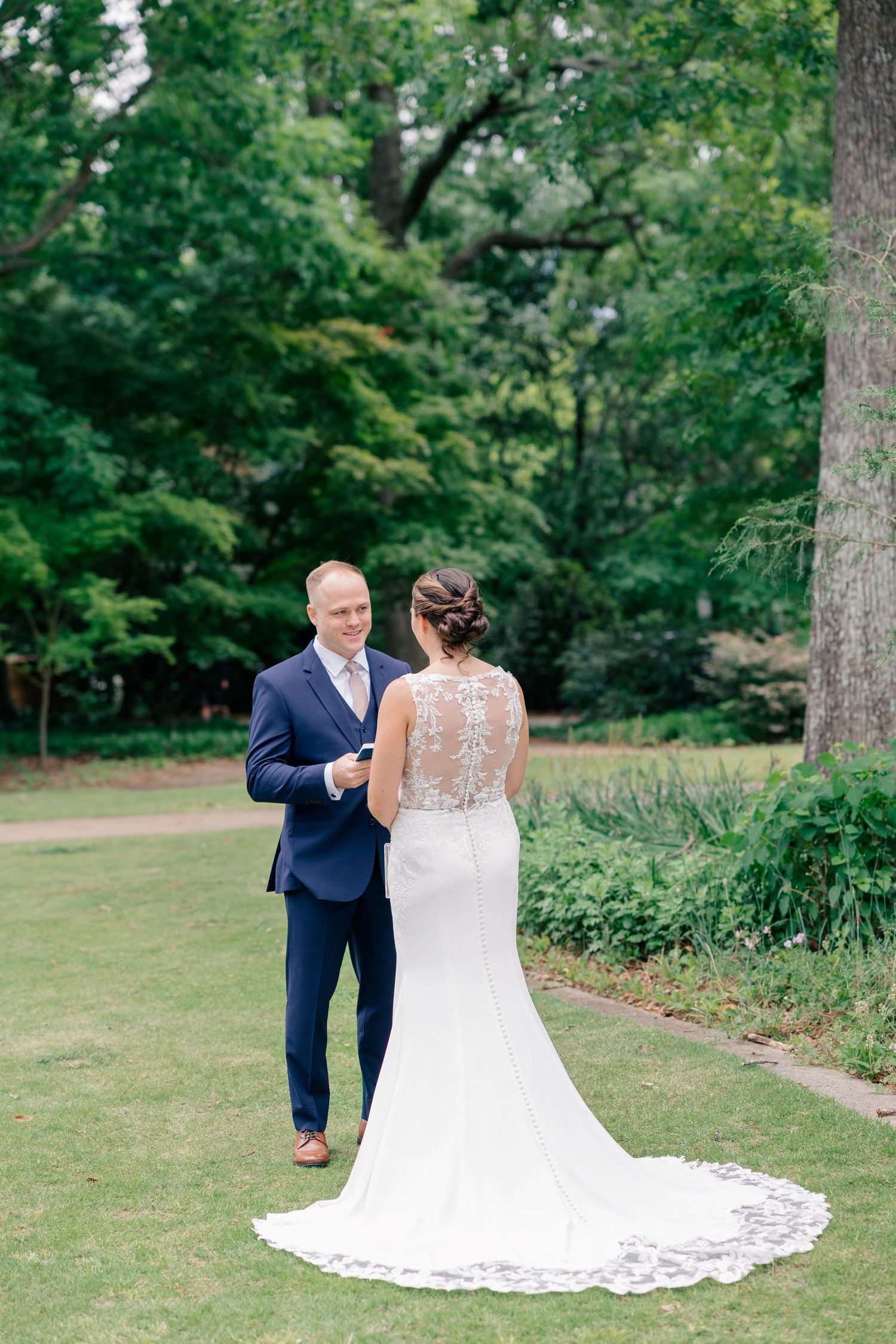 Bride in lace gown and groom in navy suit share first look moment in lush green garden.