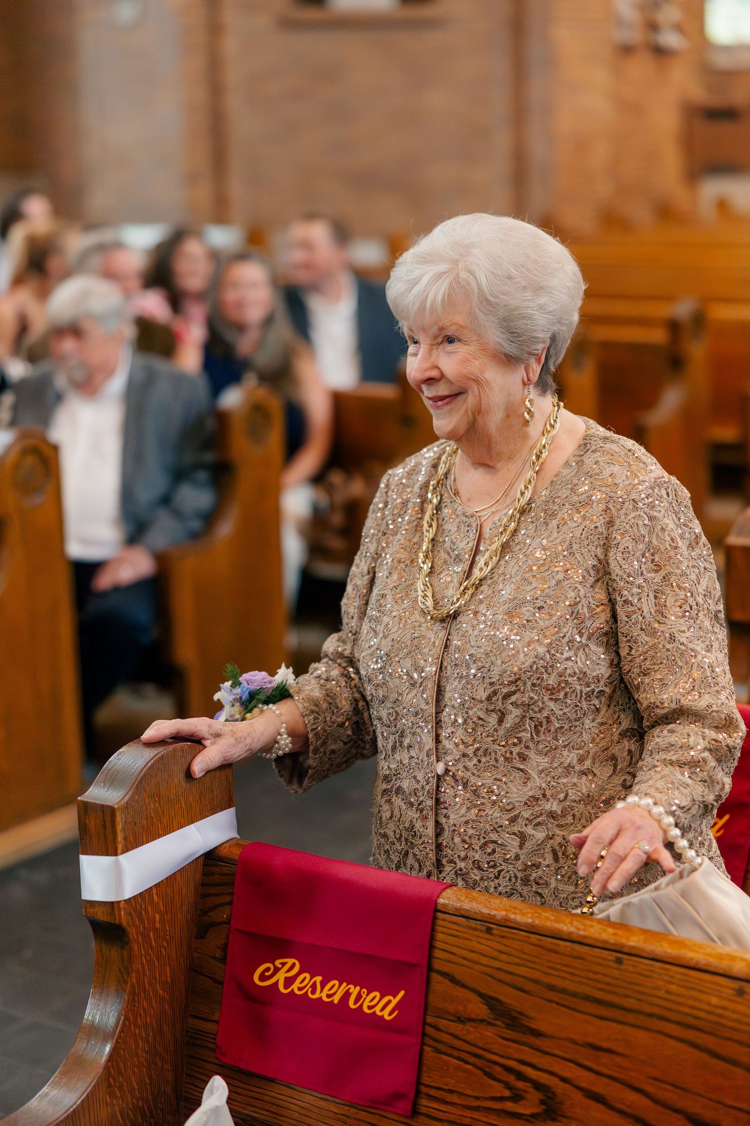 Elegant elderly woman in gold beaded dress smiles while standing at reserved church pew during wedding ceremony.