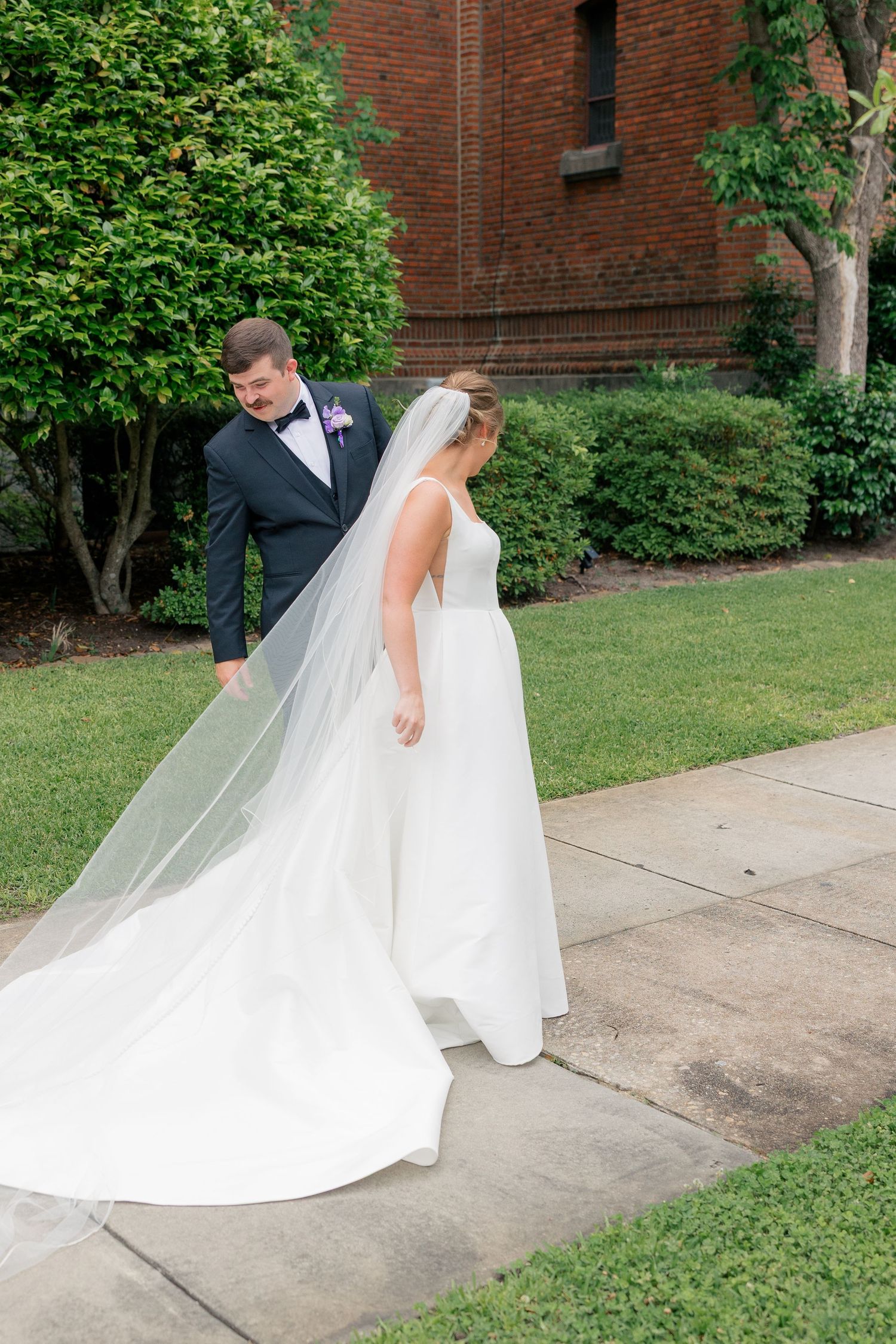 Groom in navy suit helps bride with long cathedral veil and white ball gown outside brick church.