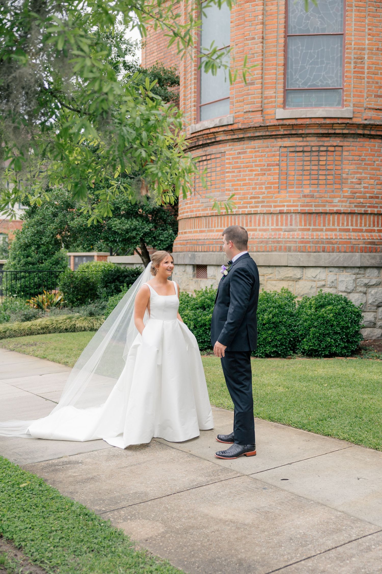 Bride and groom share a first look moment outside a historic brick church surrounded by lush greenery.
