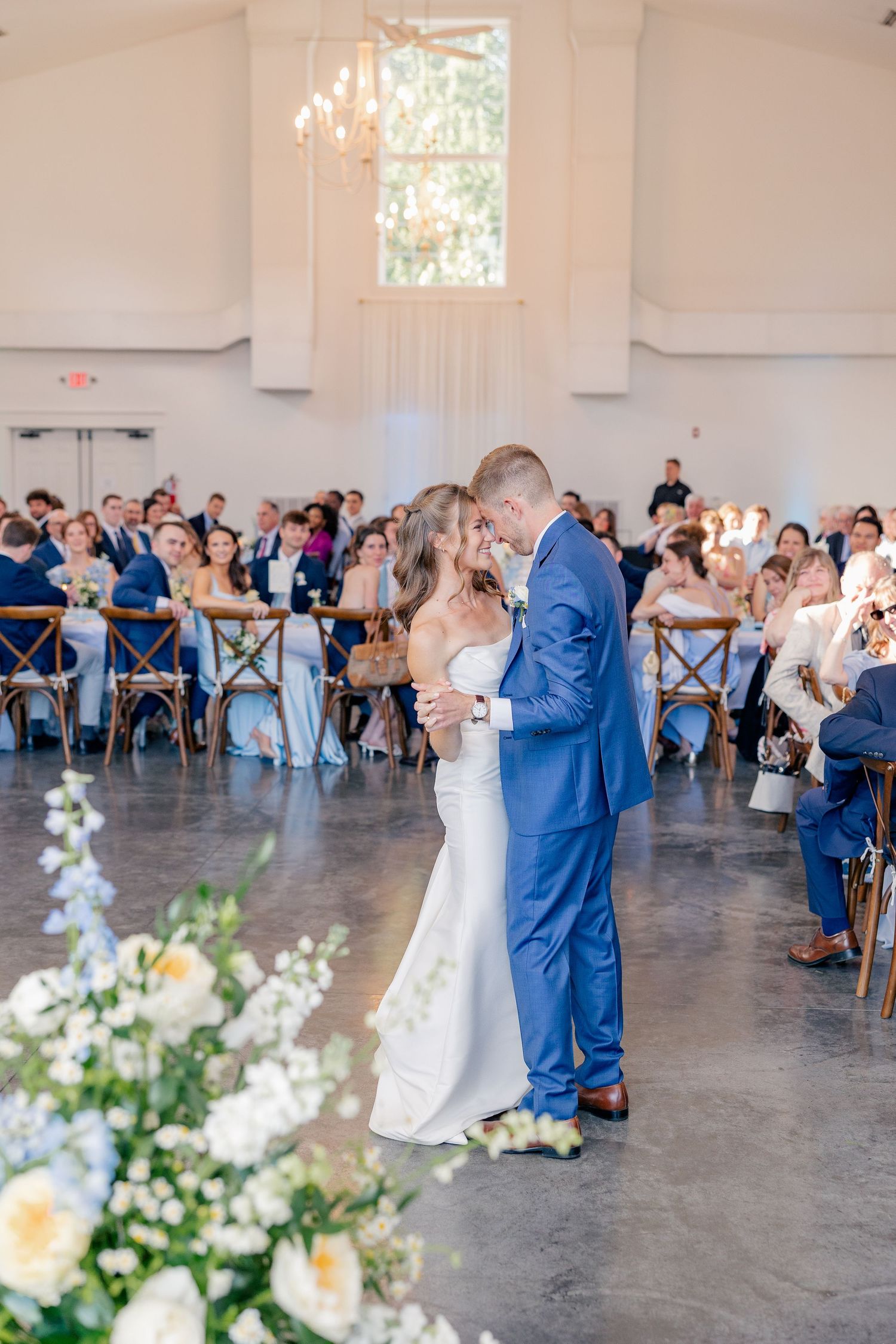 Bride and groom share first dance in elegant white reception hall with guests seated around them.