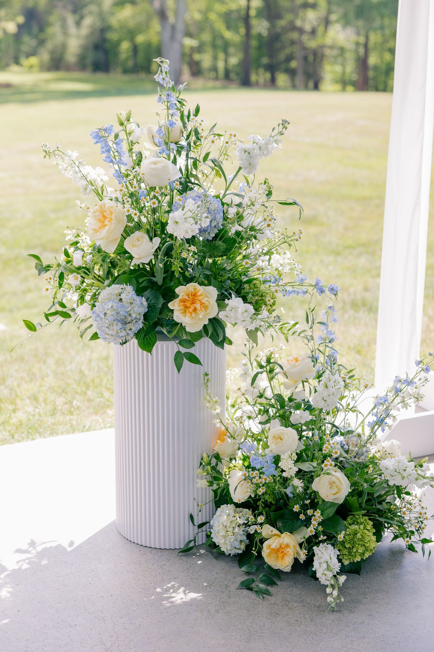 Elegant wedding floral arrangement with blue hydrangeas, yellow roses, and white blooms in a white ribbed vase outdoors.
