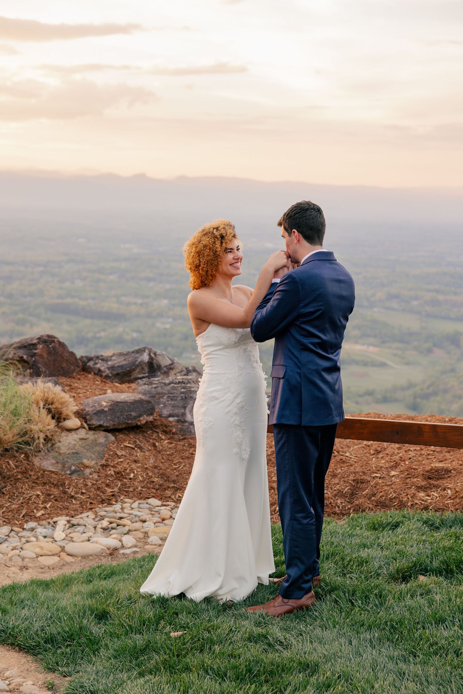 Bride in white gown and groom in navy suit share a tender moment at sunset overlook with scenic valley views.