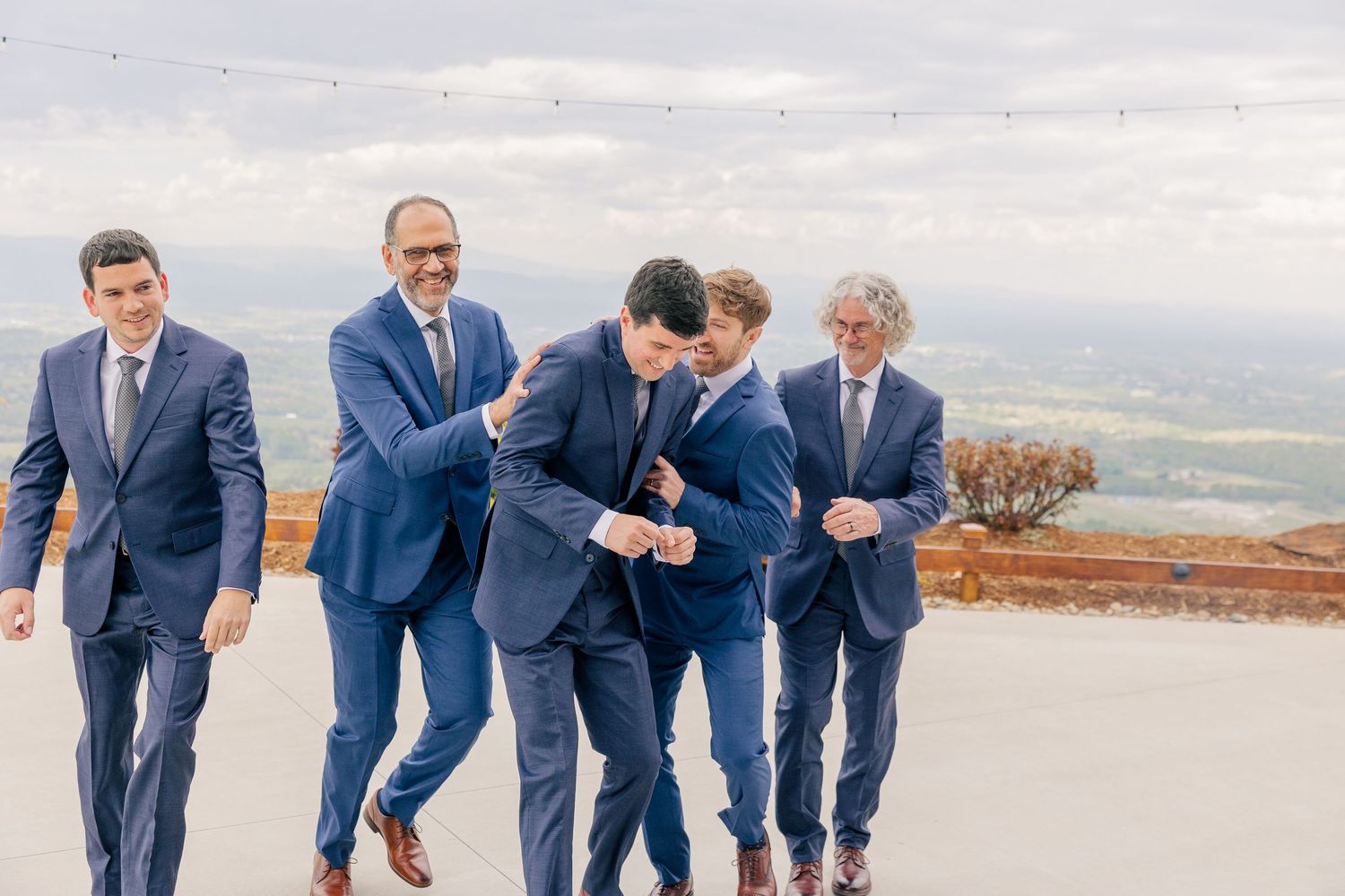 Groomsmen in navy suits laughing and joking together on outdoor terrace with scenic valley views.