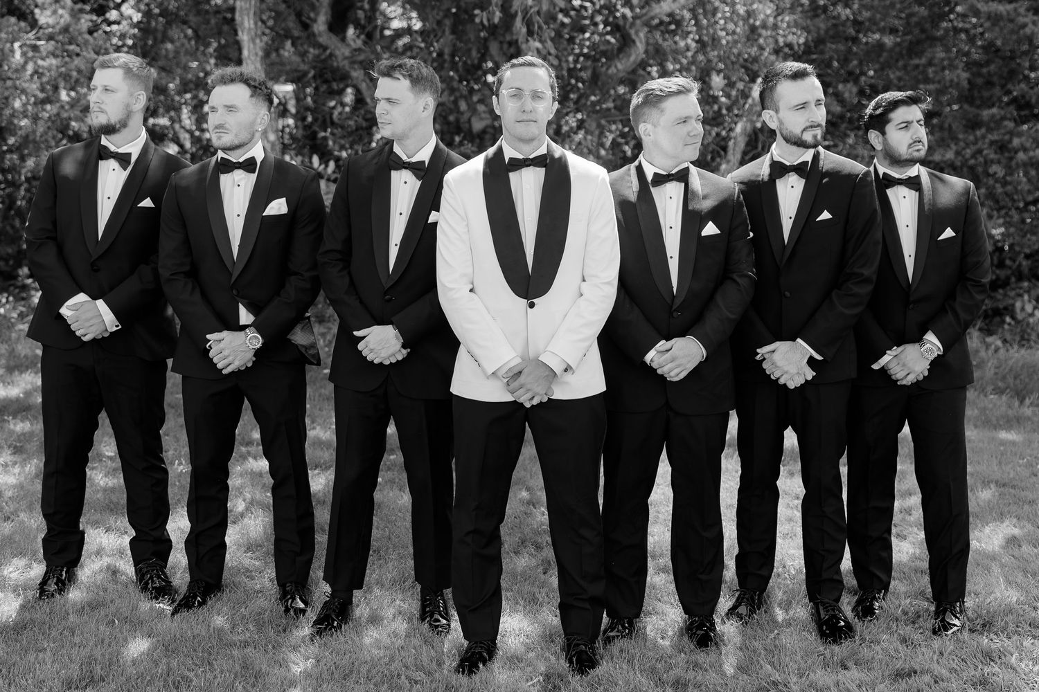 Groom in white tuxedo jacket stands with six groomsmen in black tuxedos outdoors in black and white photo.