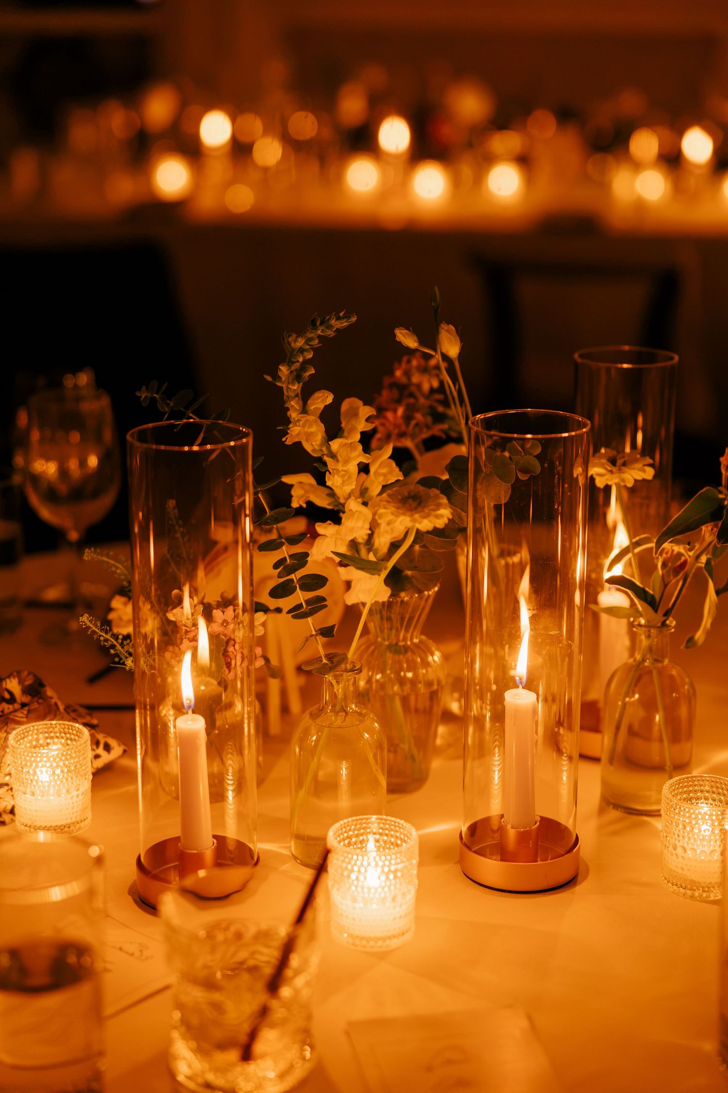 Romantic wedding table centerpiece with tall glass candle holders and small floral arrangements glowing.