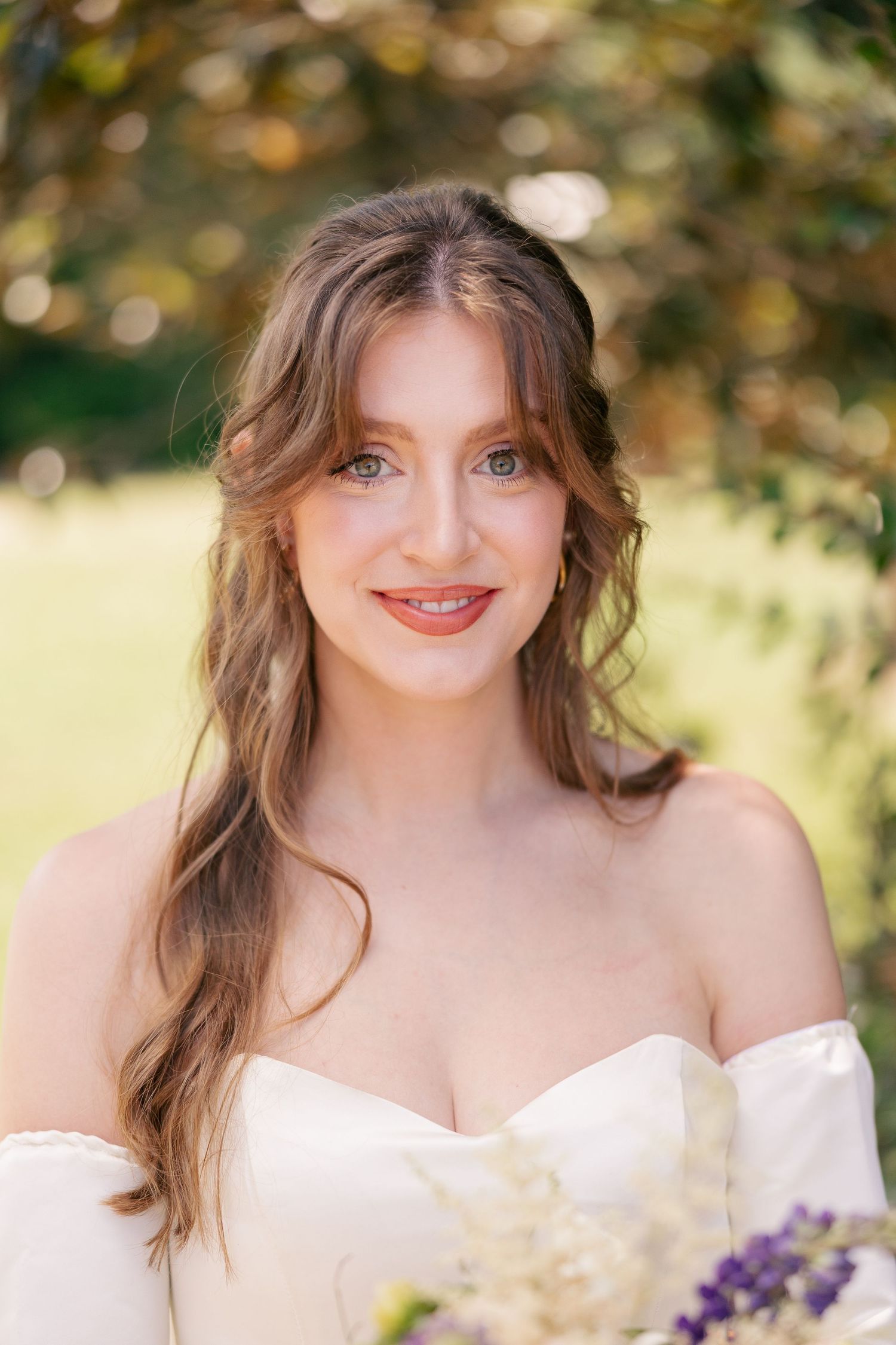 Smiling bride with wavy brown hair in an off-shoulder white gown holding wildflowers outdoors.