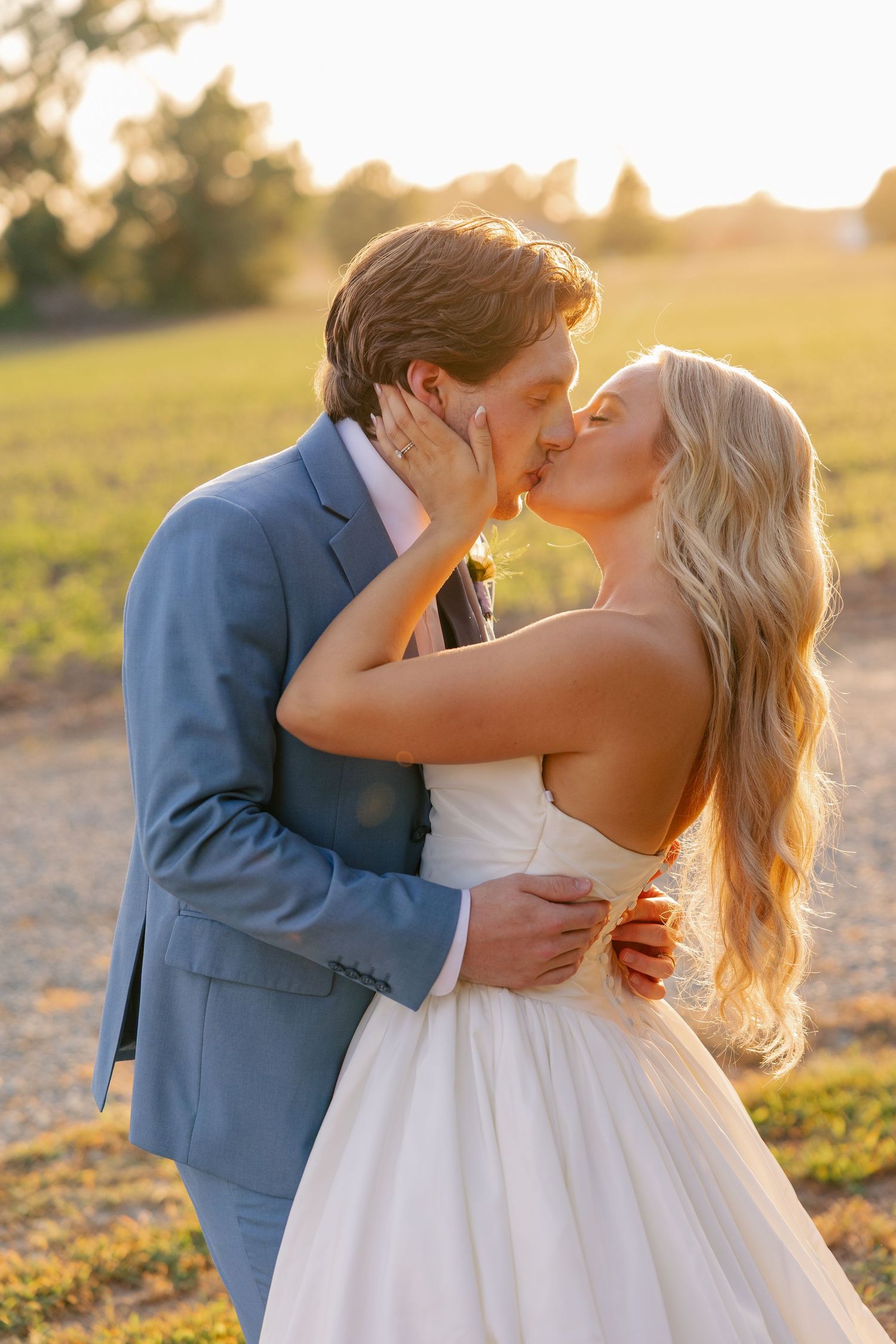 Bride and groom share passionate kiss in open field bathed in warm golden hour sunset light.
