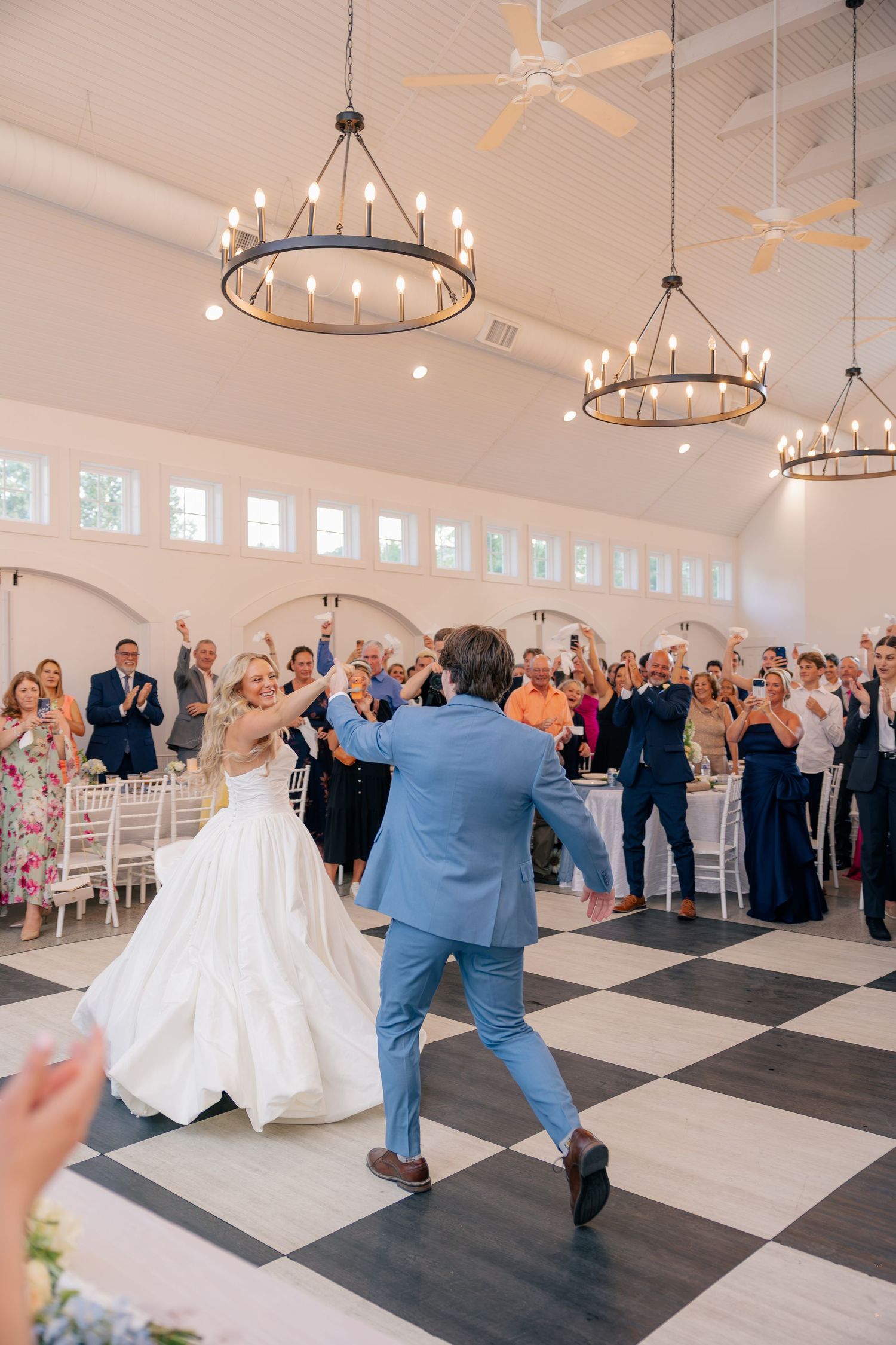 Bride and groom share energetic first dance on checkered floor in elegant white reception hall.