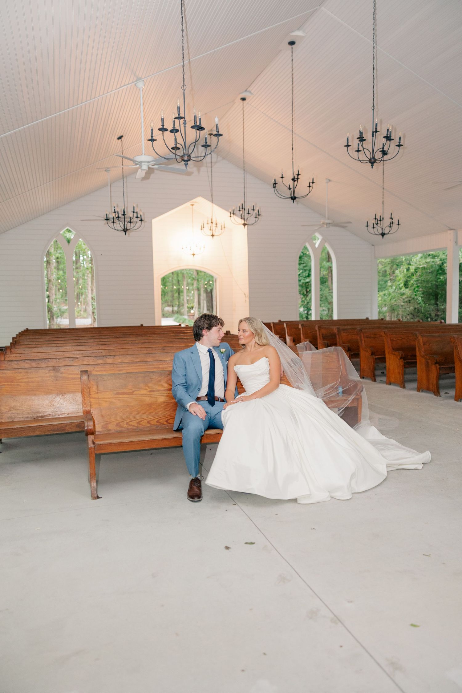 Bride and groom sitting intimately on wooden pew inside bright white chapel with arched windows.