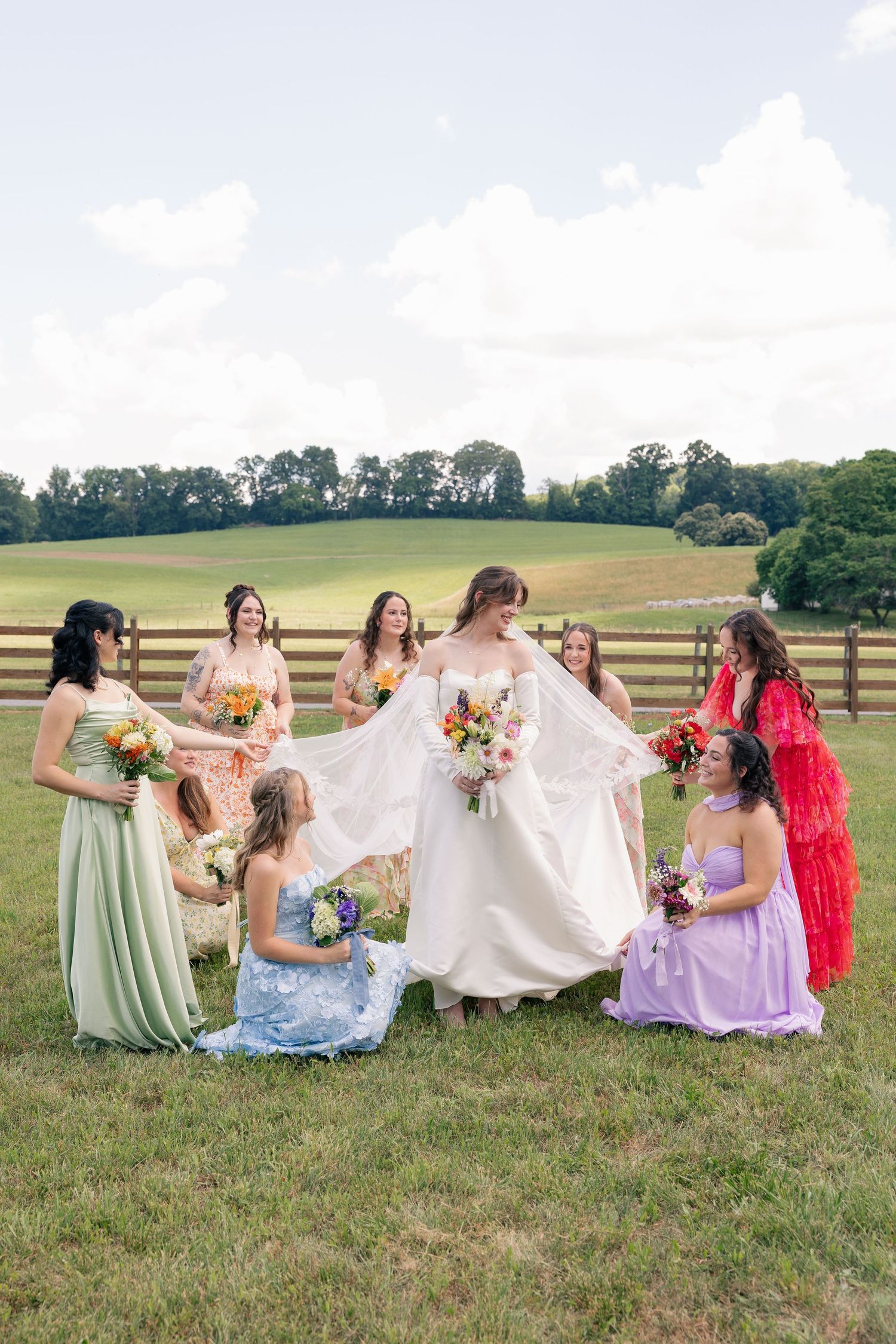 Bride surrounded by colorful mismatched bridesmaid dresses on a green farm field.