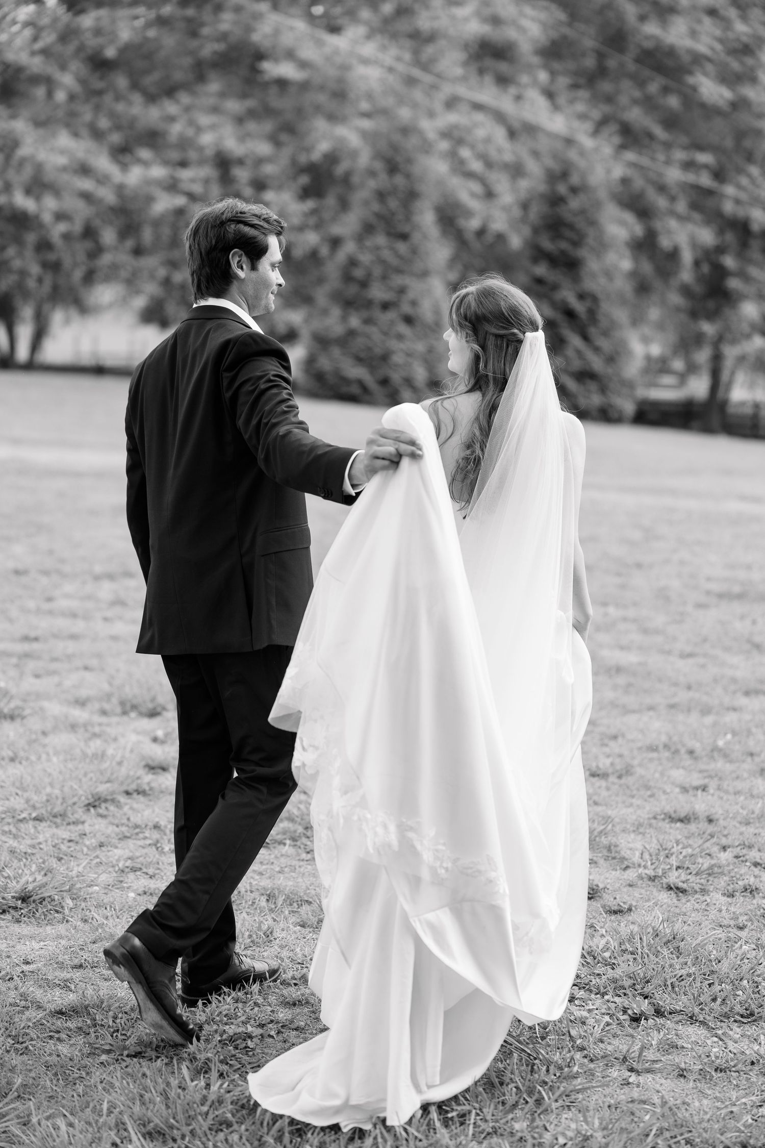 Black and white photo of groom holding bride's veil train as they walk across lawn.