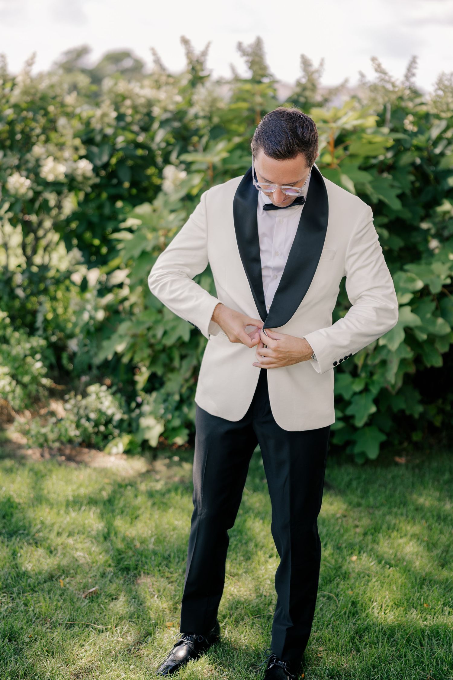 Groom in white tuxedo jacket with black lapels and bow tie buttoning his jacket outdoors near green bushes.