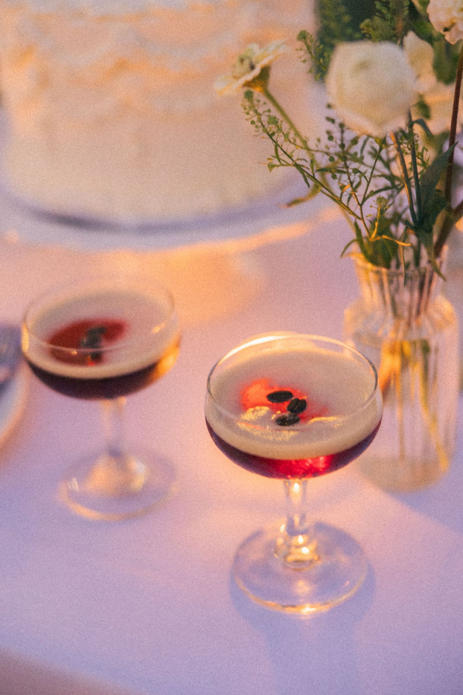 Two elegant cocktails in coupe glasses on a wedding table with white floral arrangement and tiered cake in background.