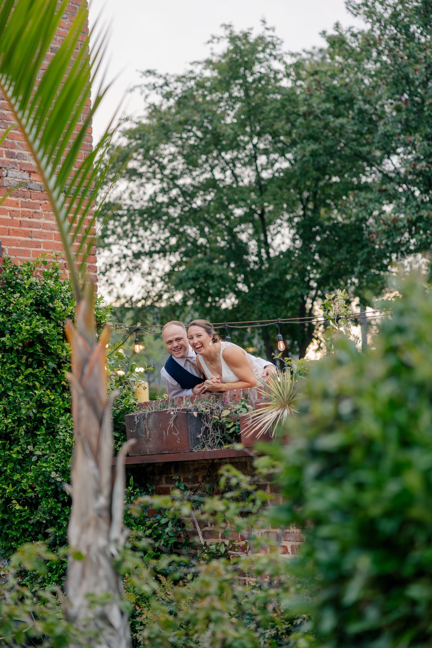 A smiling couple leans over a brick balcony surrounded by lush greenery at an outdoor wedding venue.