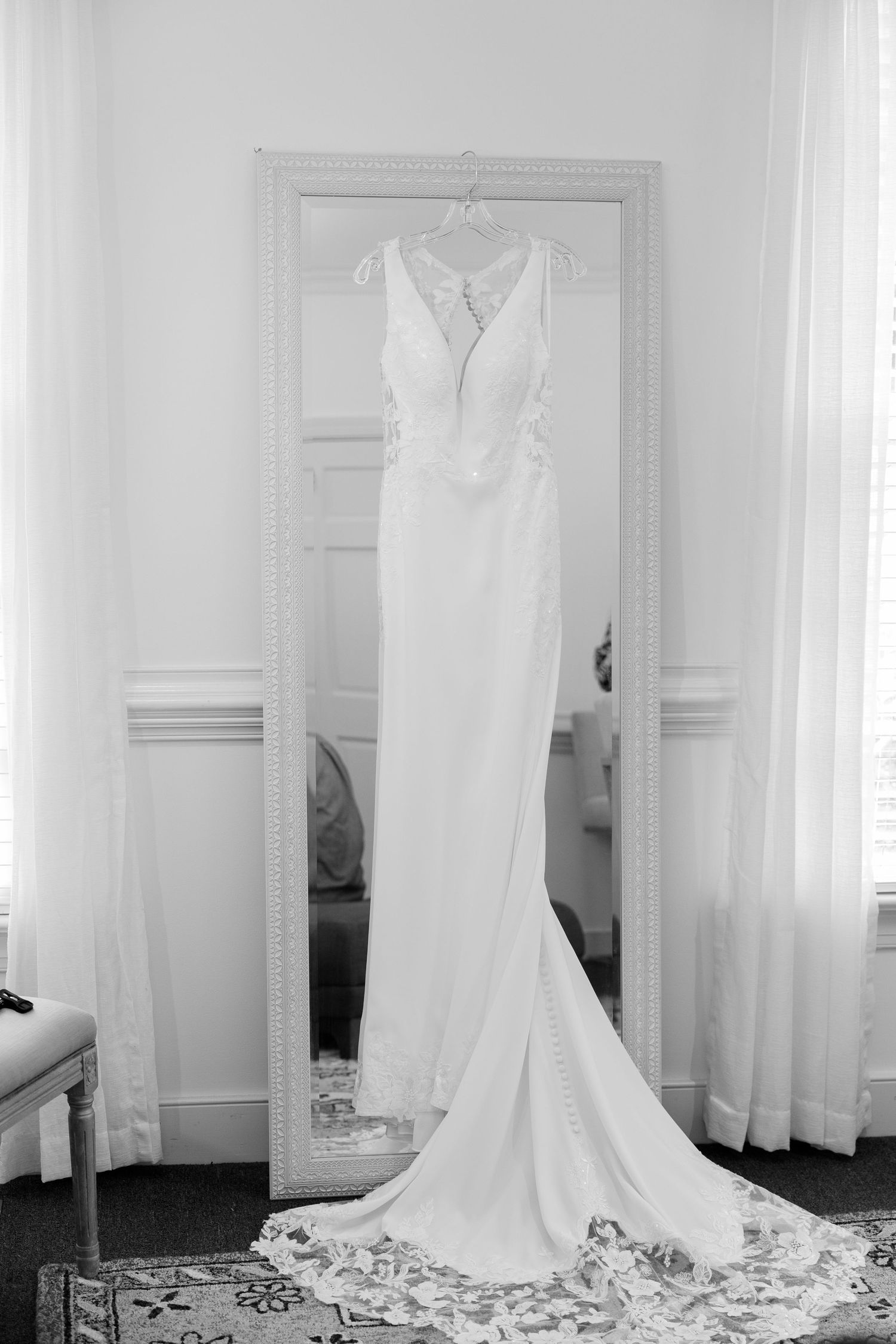 Elegant white wedding gown with lace back hanging on a mirror in a bright white room.