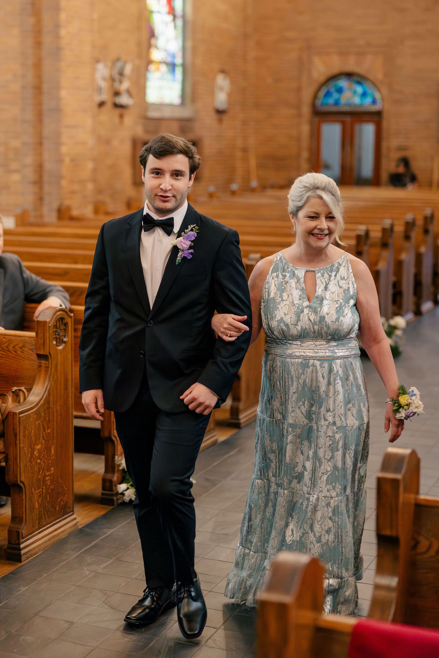 Groom in black tuxedo escorting smiling woman in blue gown down church aisle.